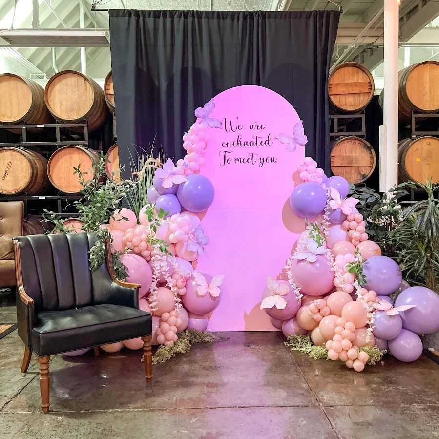A decorative backdrop with purple and pink balloons, butterflies, and greenery, featuring the quote 'We are enchanted to meet you'; located indoors with wine barrels in the background.