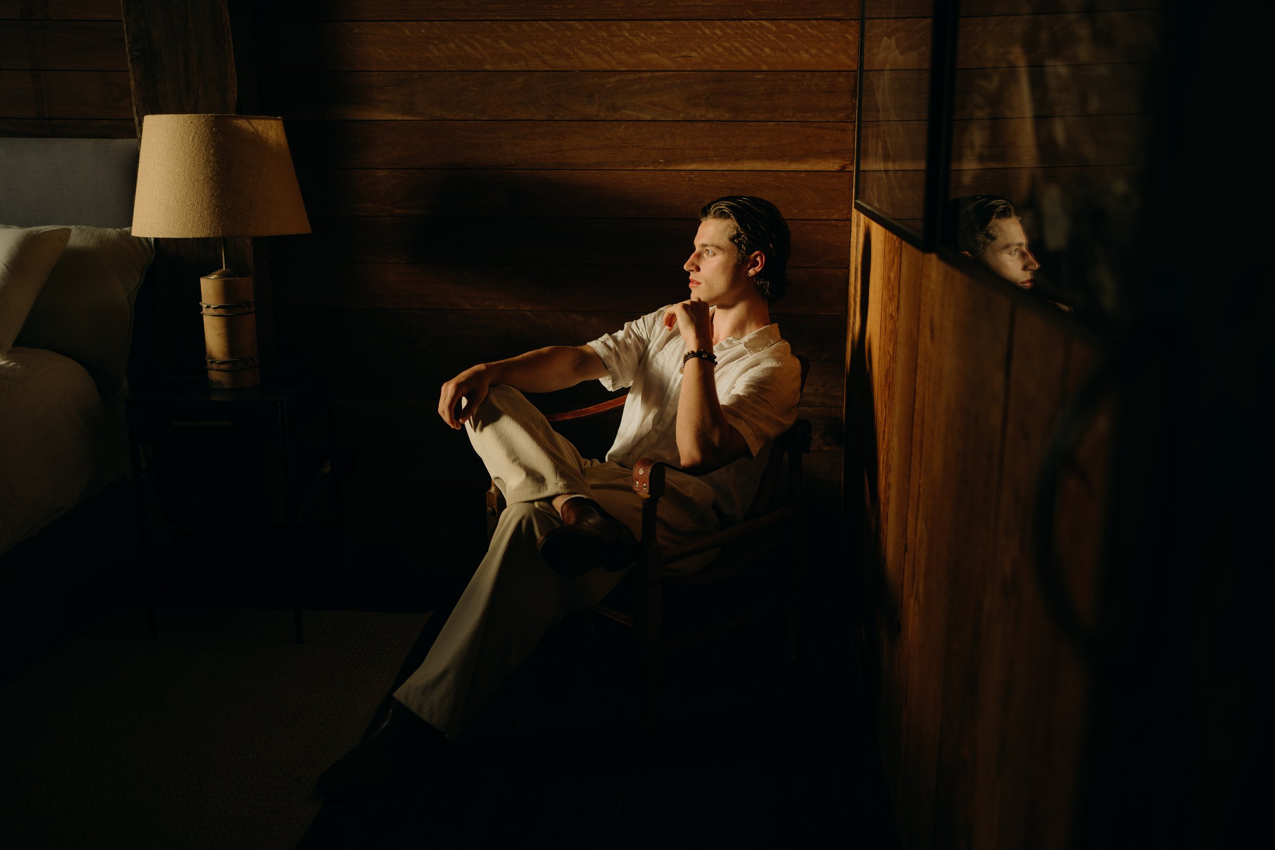 Man sitting on chair in dimly lit room, wearing a white shirt and light pants, with wooden walls and a reflection on a glass surface.