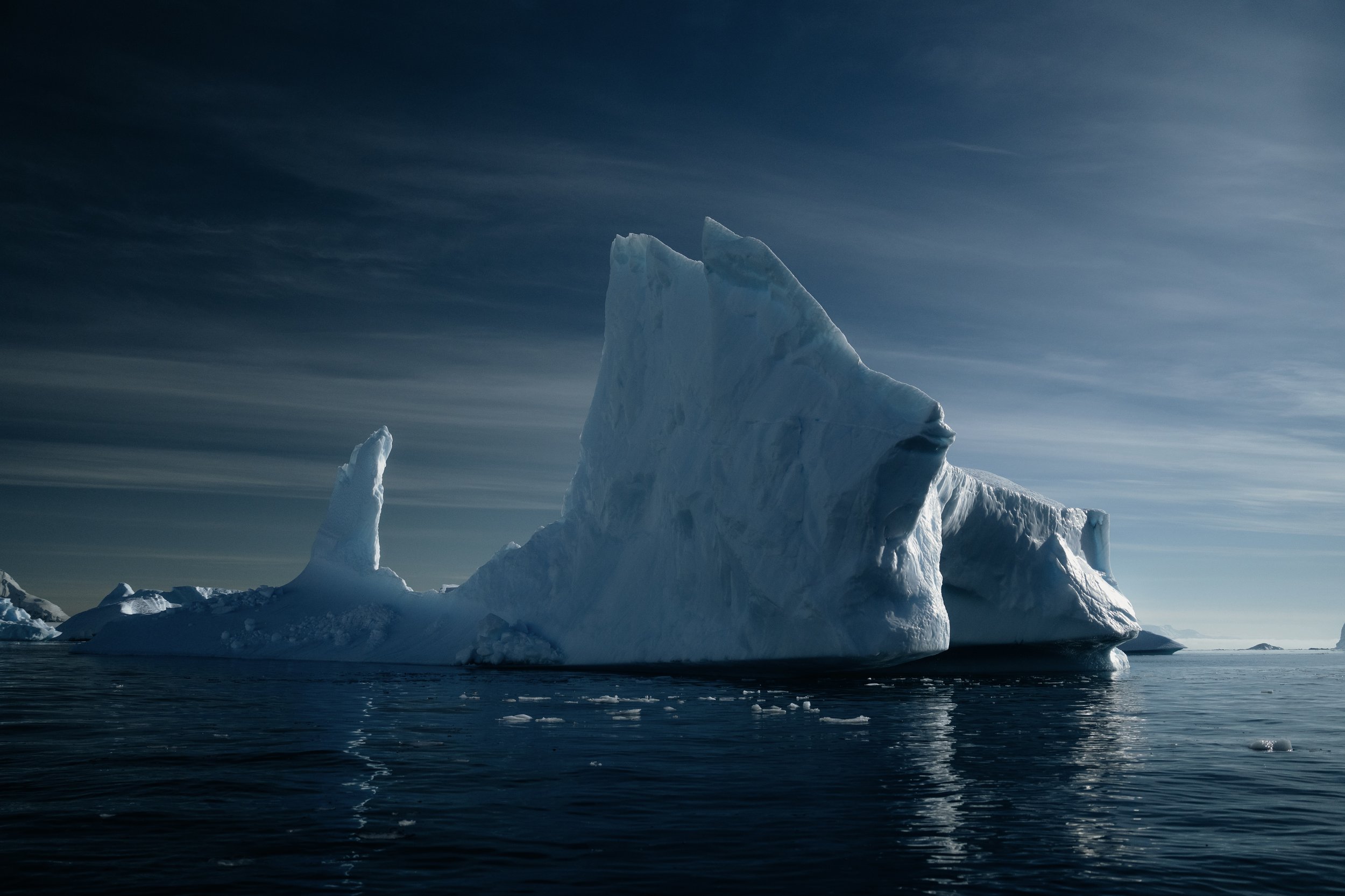 Large iceberg surrounded by calm ocean water under a cloudy sky, reflecting the ice and light.