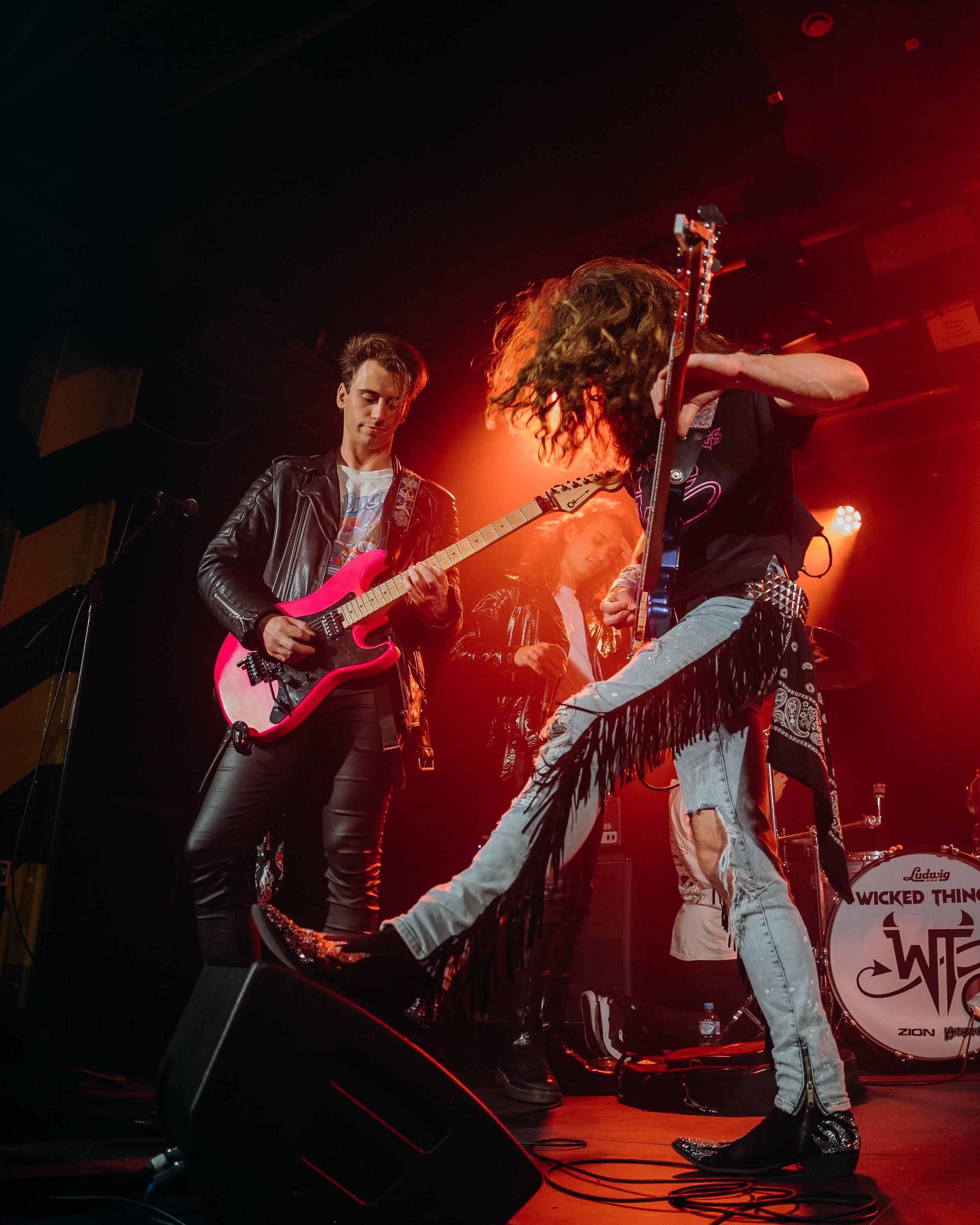 Rock band performing on stage with guitarist playing pink guitar, another guitarist jumping, and drums in background under red lighting.