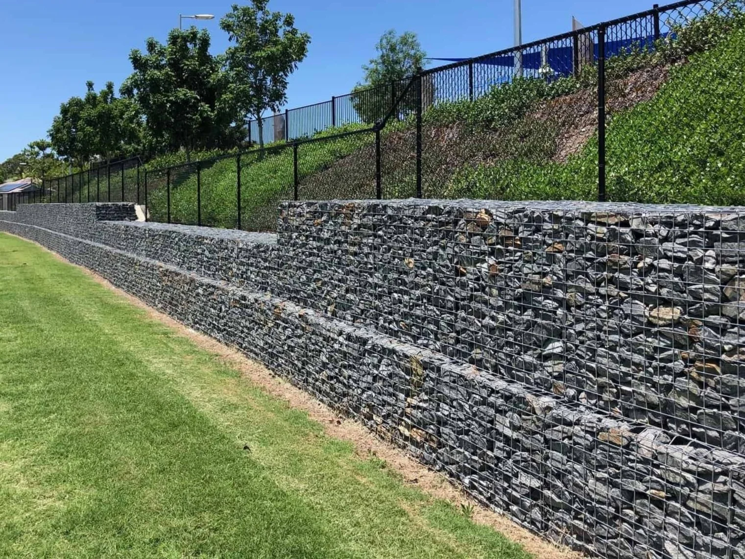 A rock-filled gabion retaining wall along a grassy area with trees and a fence on a hillside under a sunny, clear blue sky.