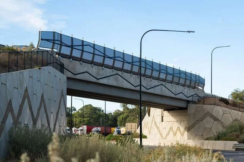 A modern overpass bridge with geometric patterned concrete supports and a glass barrier, with vehicles passing underneath and a clear blue sky in the background.