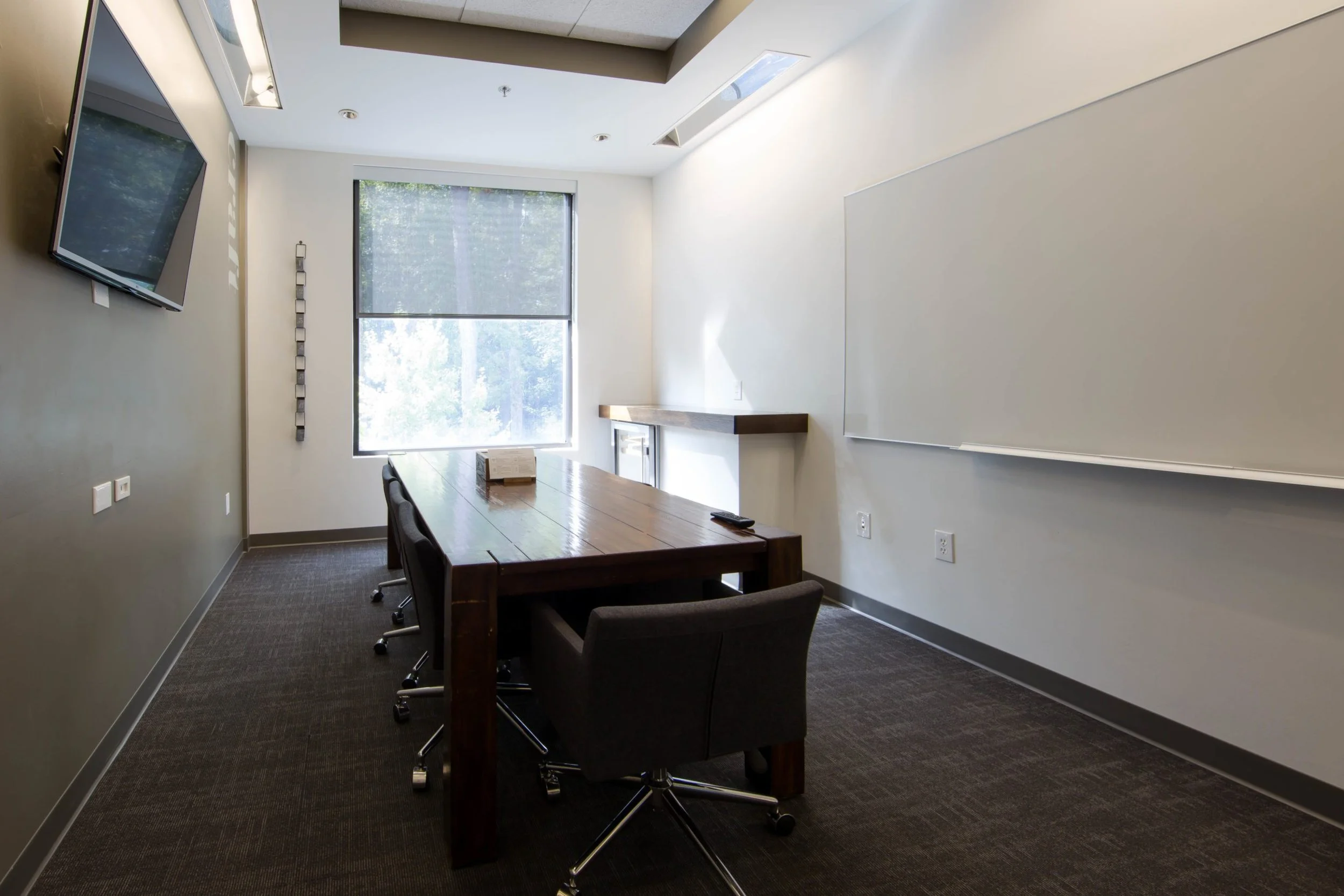 Conference room with a large window, wooden table, black rolling chairs, whiteboard, and a wall-mounted TV.