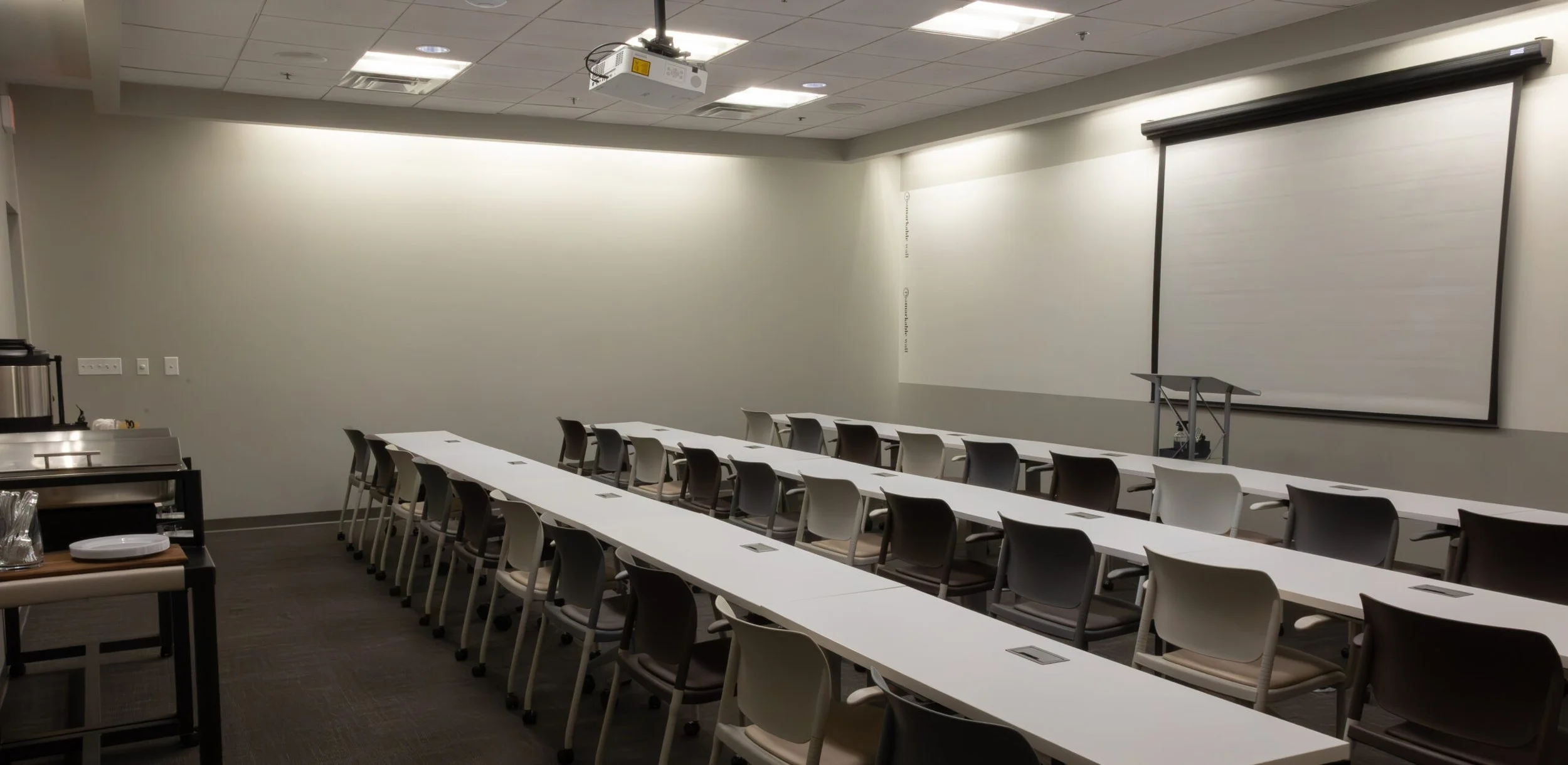 An empty conference room with white tables and gray and white chairs, a large white wall-mounted projection screen, and a ceiling-mounted projector. There is a small table with coffee supplies on the left side.