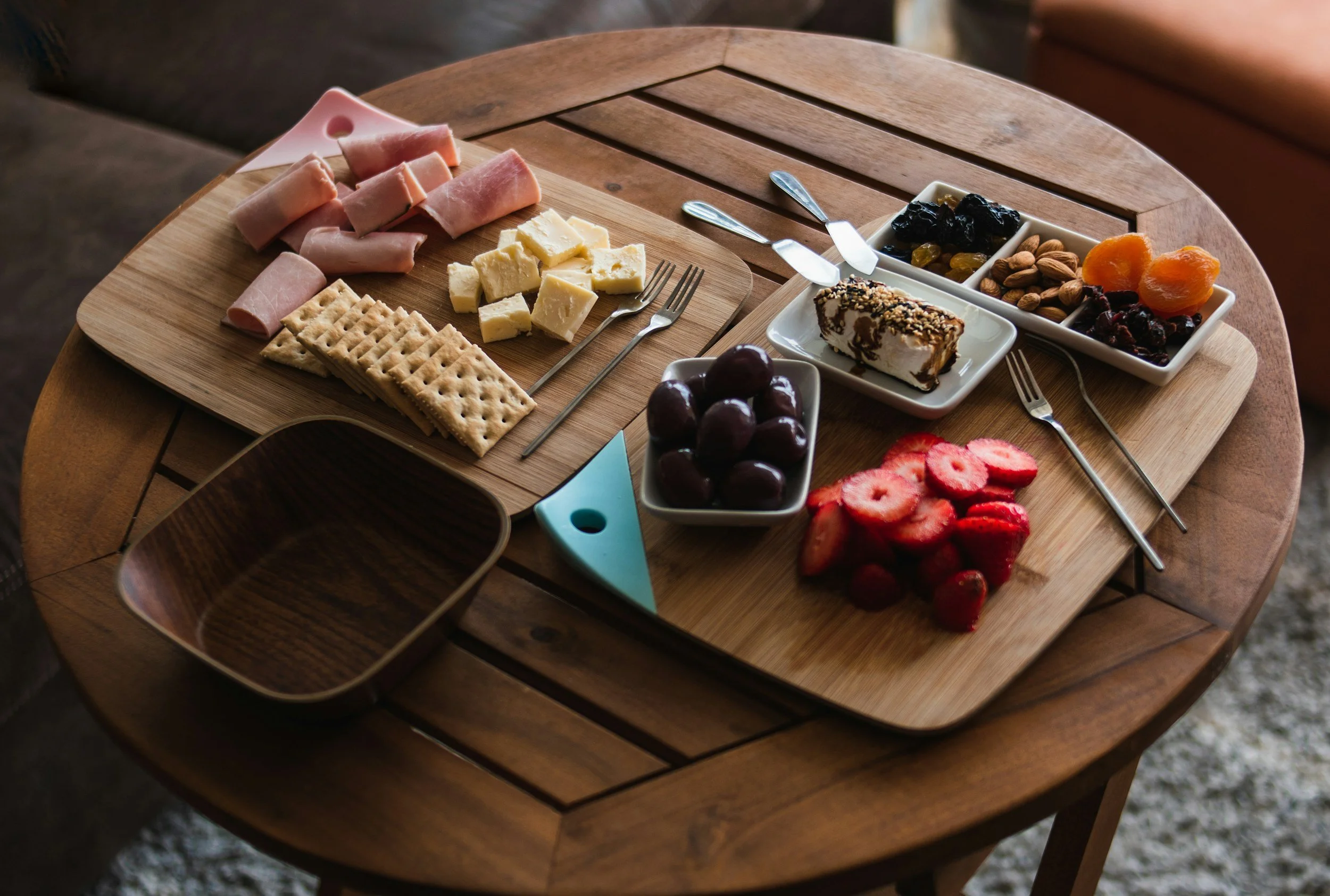 Displaying different types of snacks such as ham, cheese, crackers, olives, strawberries, brie, raisins, almonds, and dried apricots on wooden plates and bowls sitting on a wooden round side table.