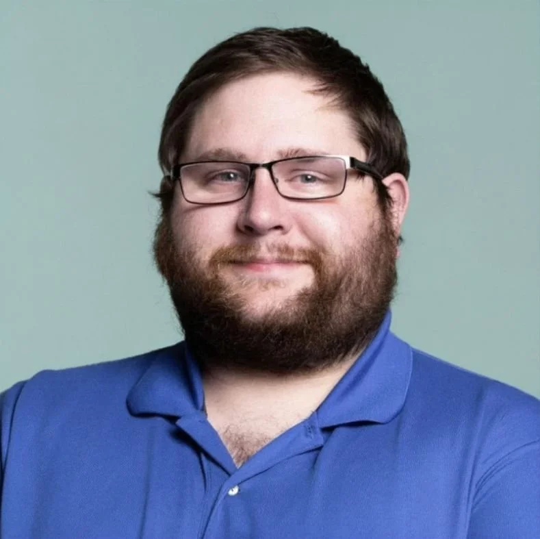 A man with glasses, a beard, and short brown hair wearing a blue polo shirt, smiling, with a plain light-colored background.