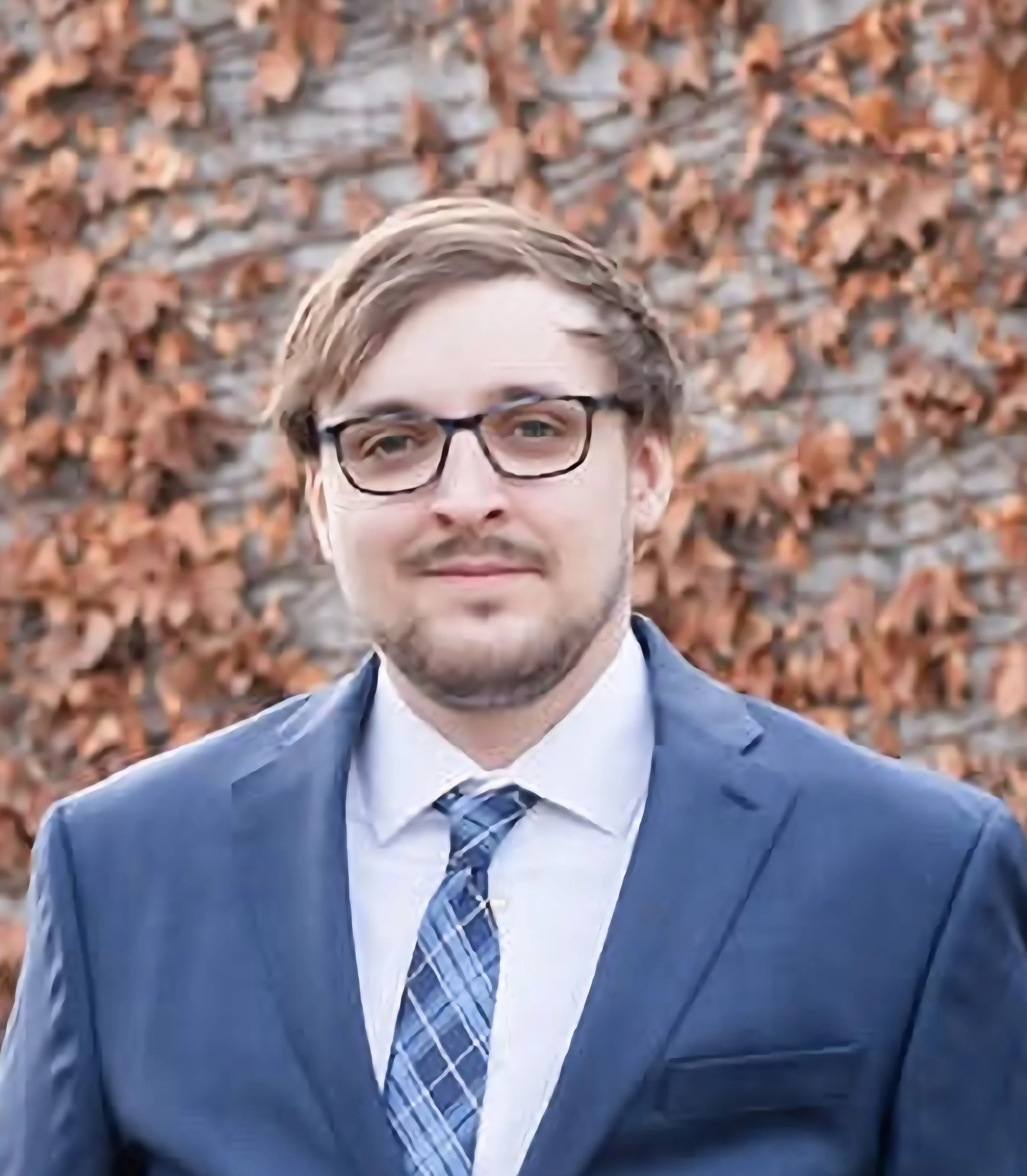 A man with glasses wearing a suit and tie, standing outdoors with orange autumn leaves in the background.