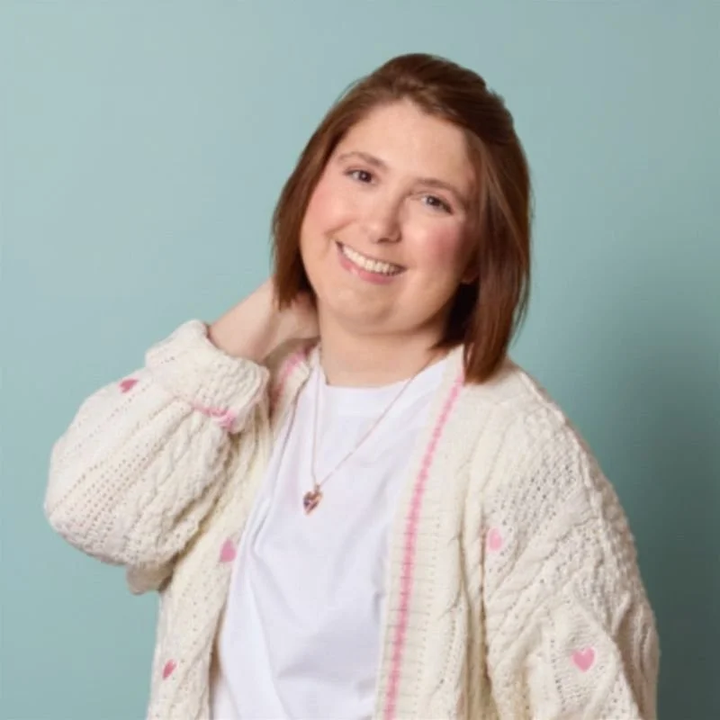 A young woman with shoulder-length brown hair, smiling, wearing a white shirt, a cream-colored cardigan with pink hearts, and a heart-shaped necklace, posing against a light blue background.