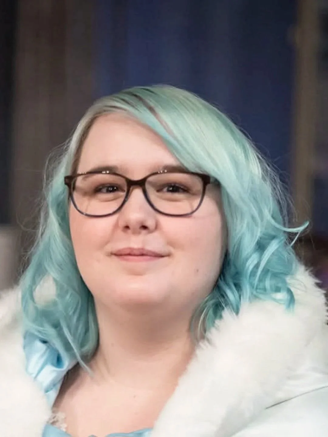 Close-up of a woman with light blue and green wavy hair, wearing black glasses and a white furry coat, smiling gently.
