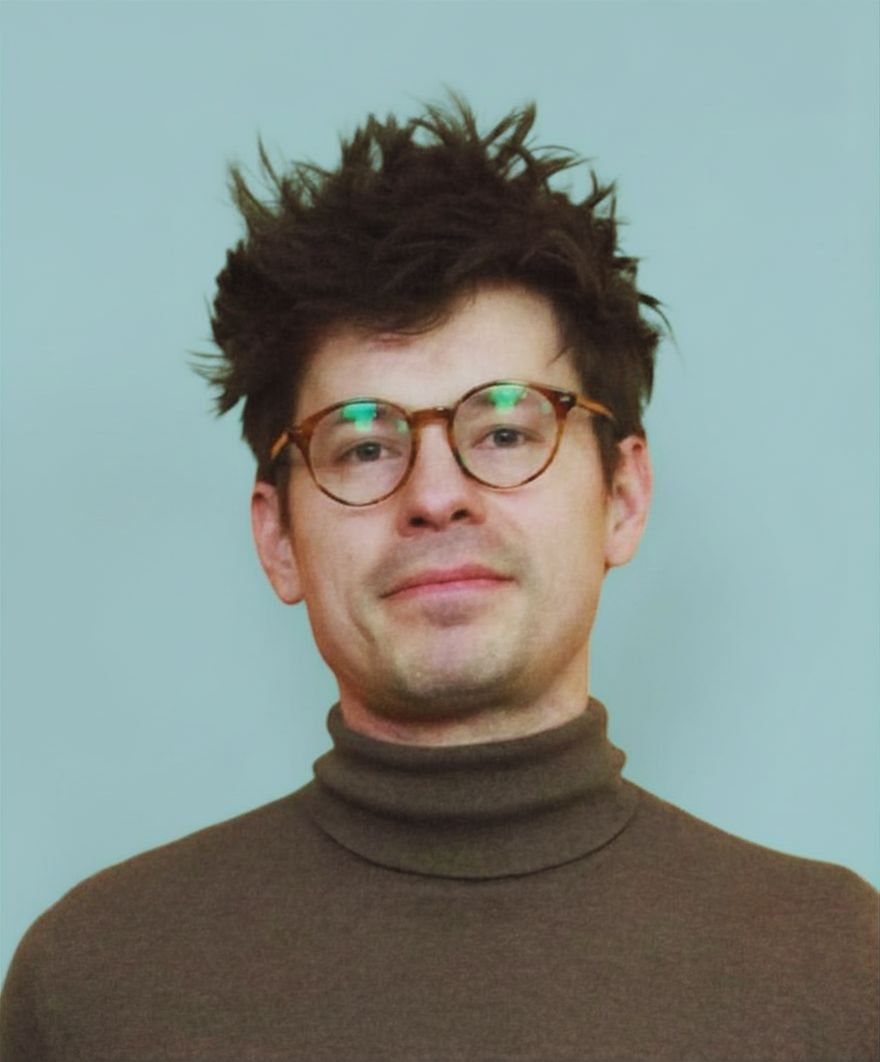 A man with messy dark hair, wearing glasses with brown frames, and a brown turtleneck sweater, standing against a light blue background.