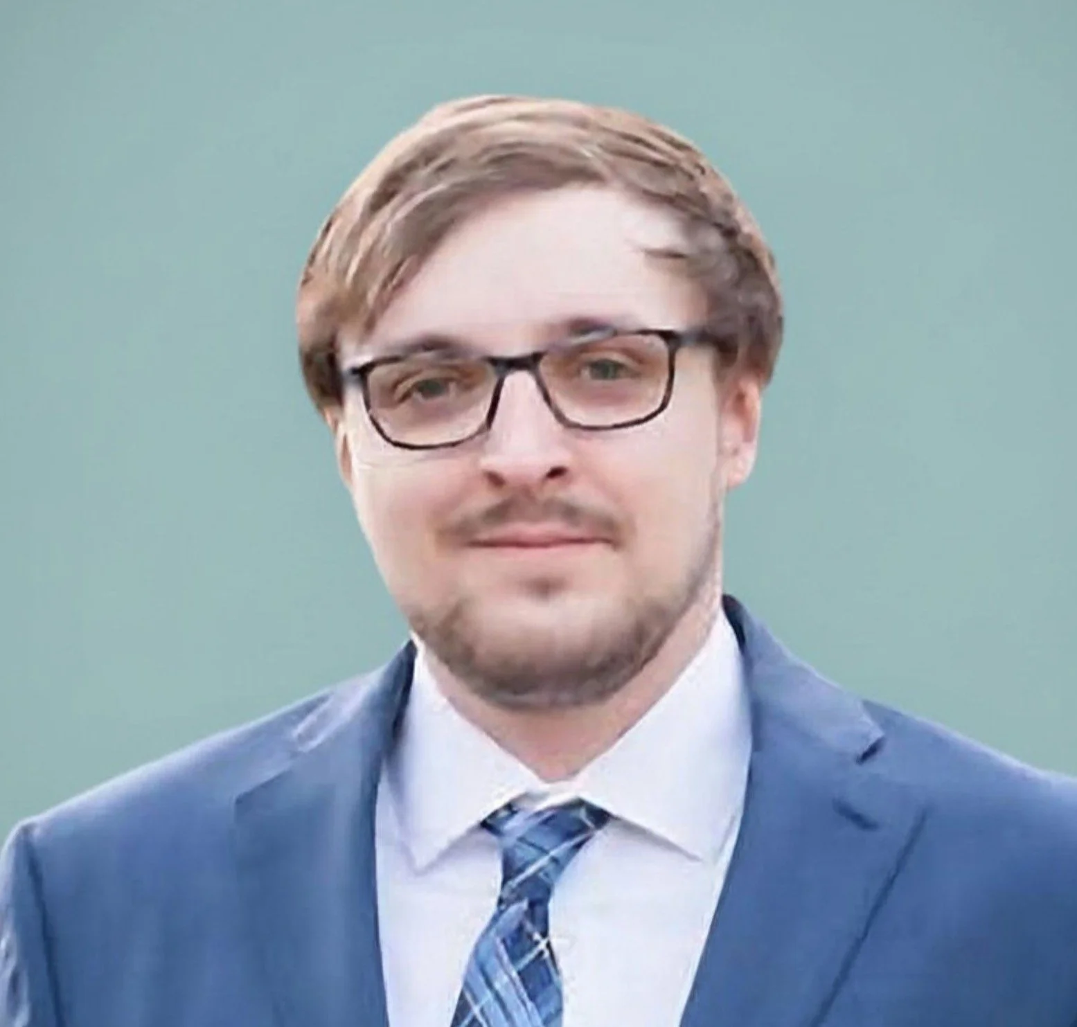 A man in a blue suit, white shirt, and patterned tie, wearing glasses, standing outdoors against a blurred green background.