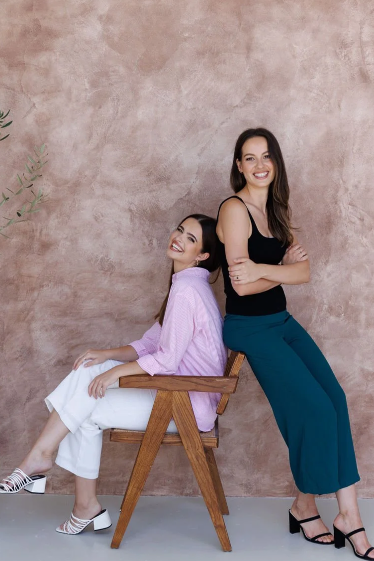 Two women smiling, one sitting on a wooden chair wearing a pink striped shirt and white pants, the other standing leaning against the chair wearing a black top and teal pants, against a textured pink wall.