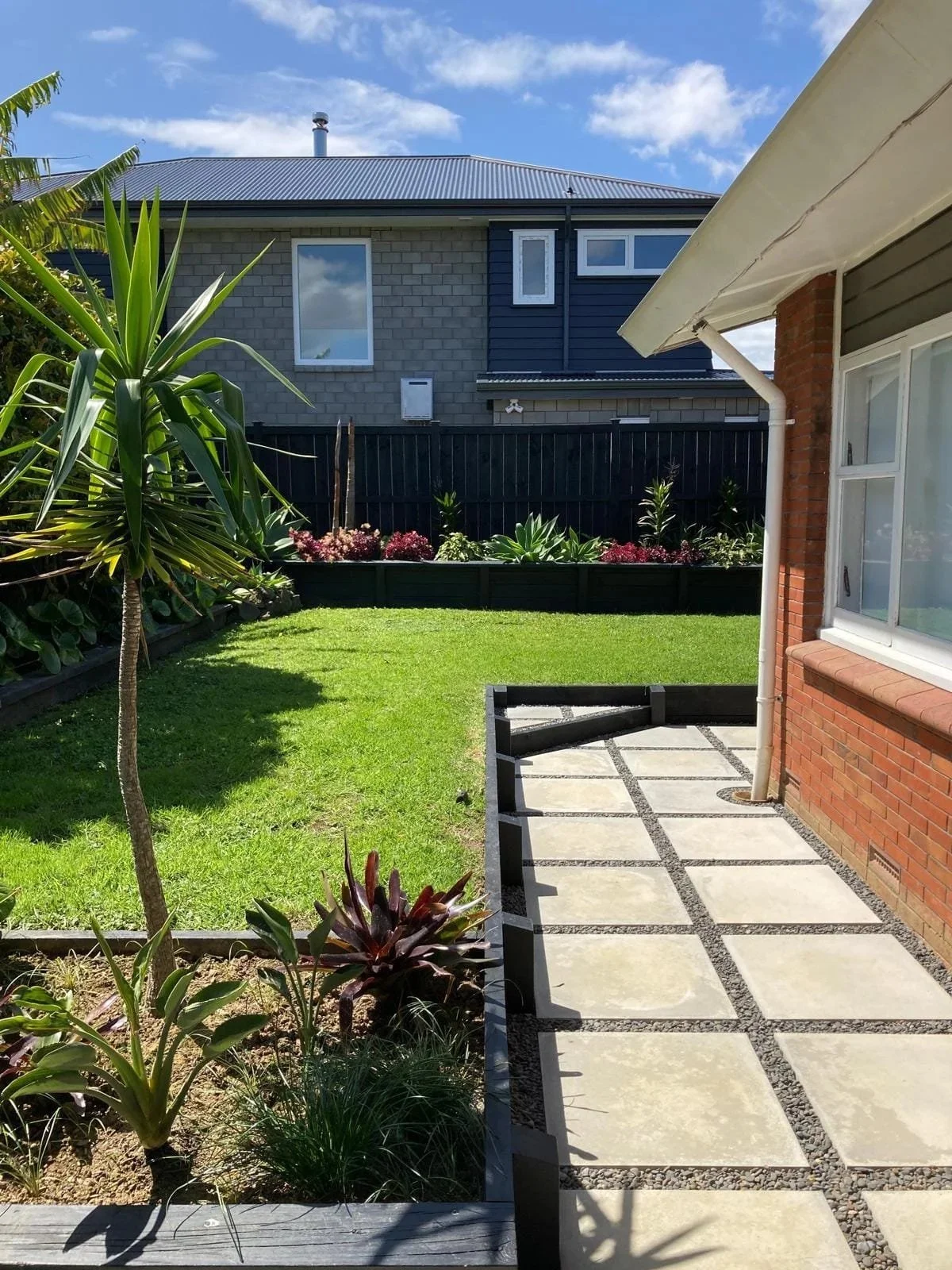 A backyard with a small grassy lawn, paved stone walkway, and various plants and shrubs, with a house in the background under a blue sky with some clouds.