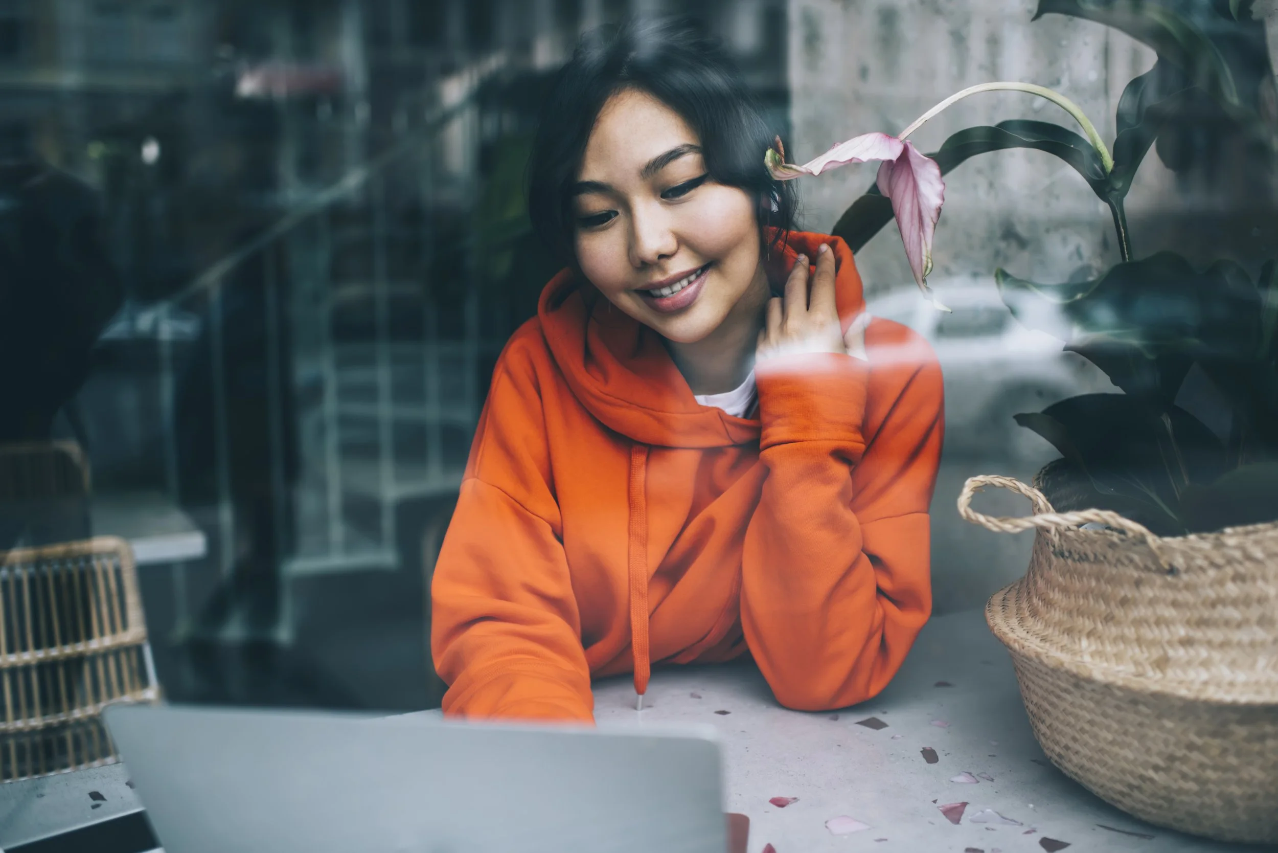 Young woman in orange hoodie smiling while looking at a laptop, sitting by a window with a pink flower and a woven basket nearby.