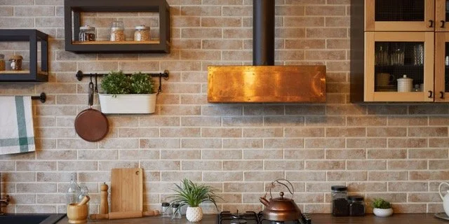 Kitchen with brick wall, wooden cabinets, hanging shelves, and various kitchen items, including a green plant and a copper range hood.