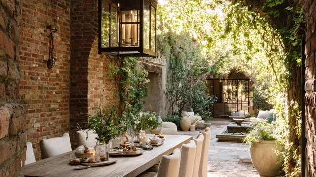 Outdoor dining area with a wooden table set with plates, cups, and candles, surrounded by potted plants, brick walls, and hanging lanterns, in a lush garden setting.