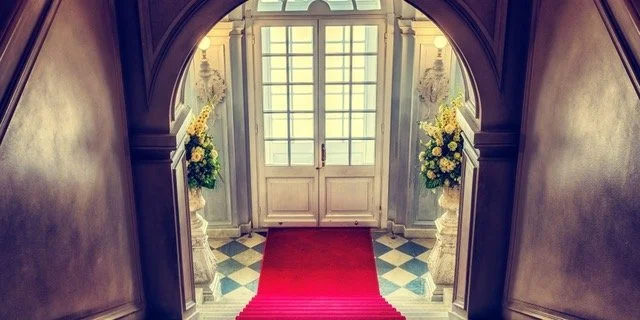 Entrance hallway with double doors, a red carpet, and flower arrangements on pedestal stands under an arched doorway.