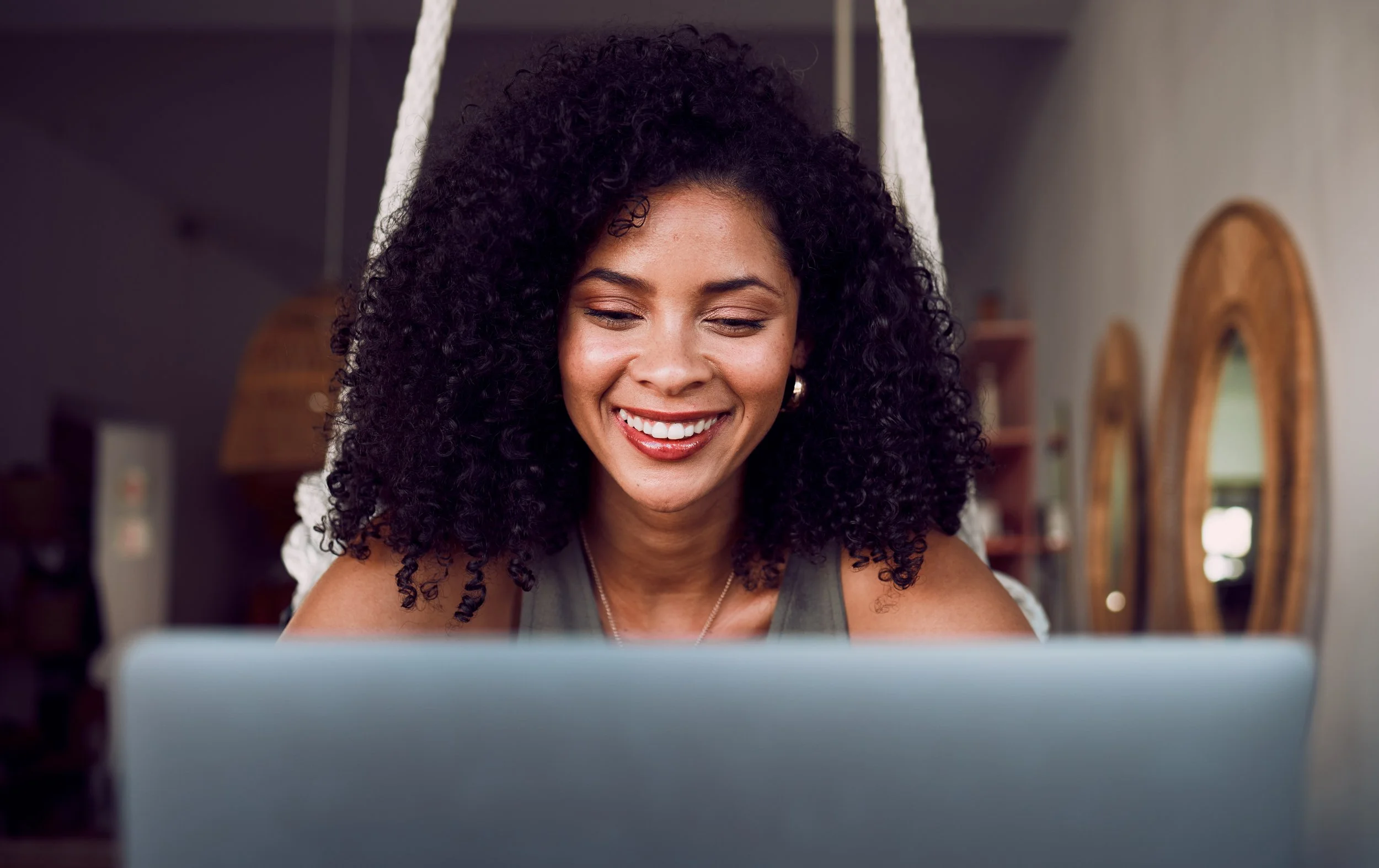 A woman with curly black hair smiling while looking at her laptop screen.