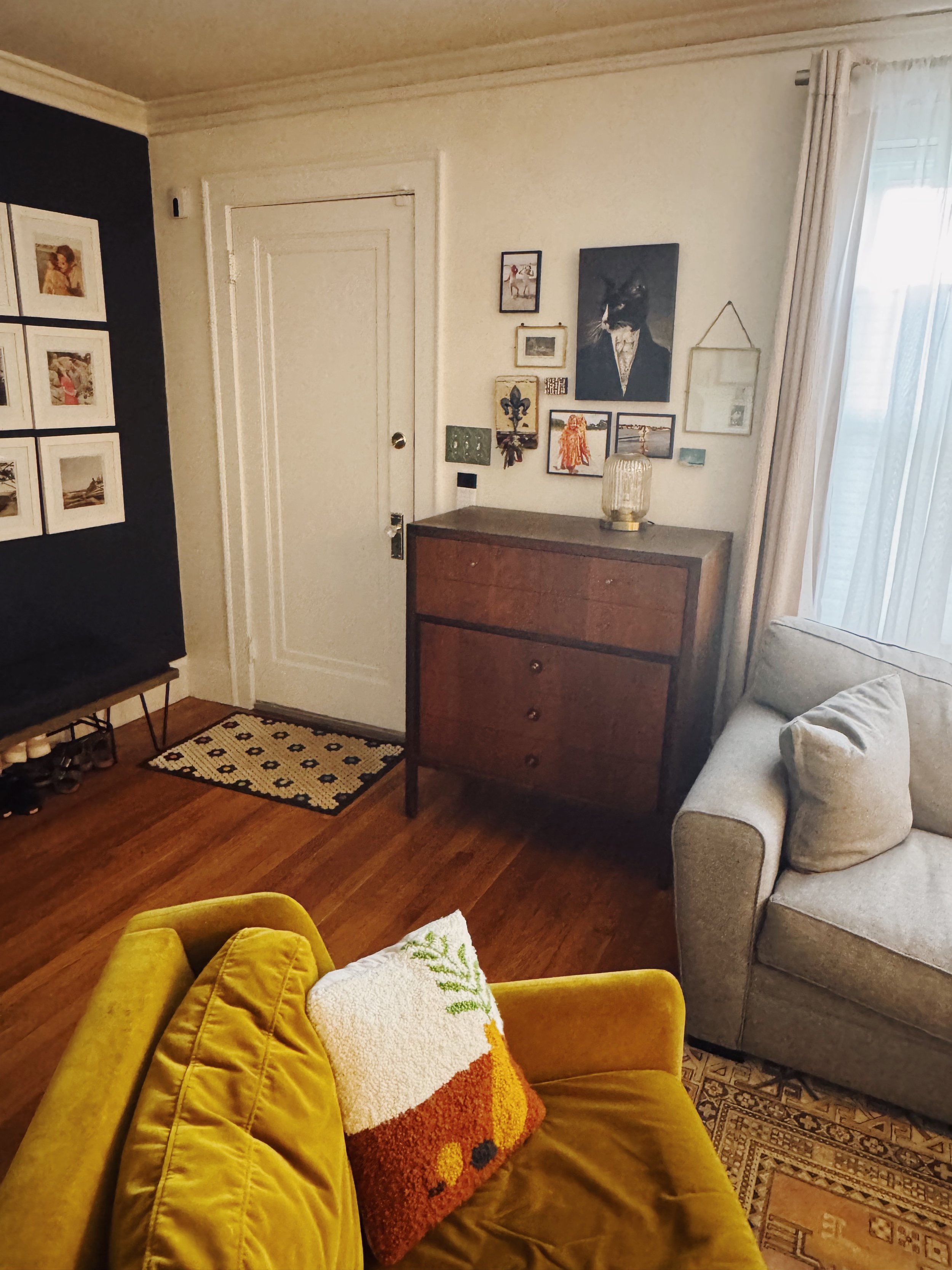 Living room corner with a yellow velvet couch, colorful throw pillow, vintage dresser, wall art, and a window with white curtains.