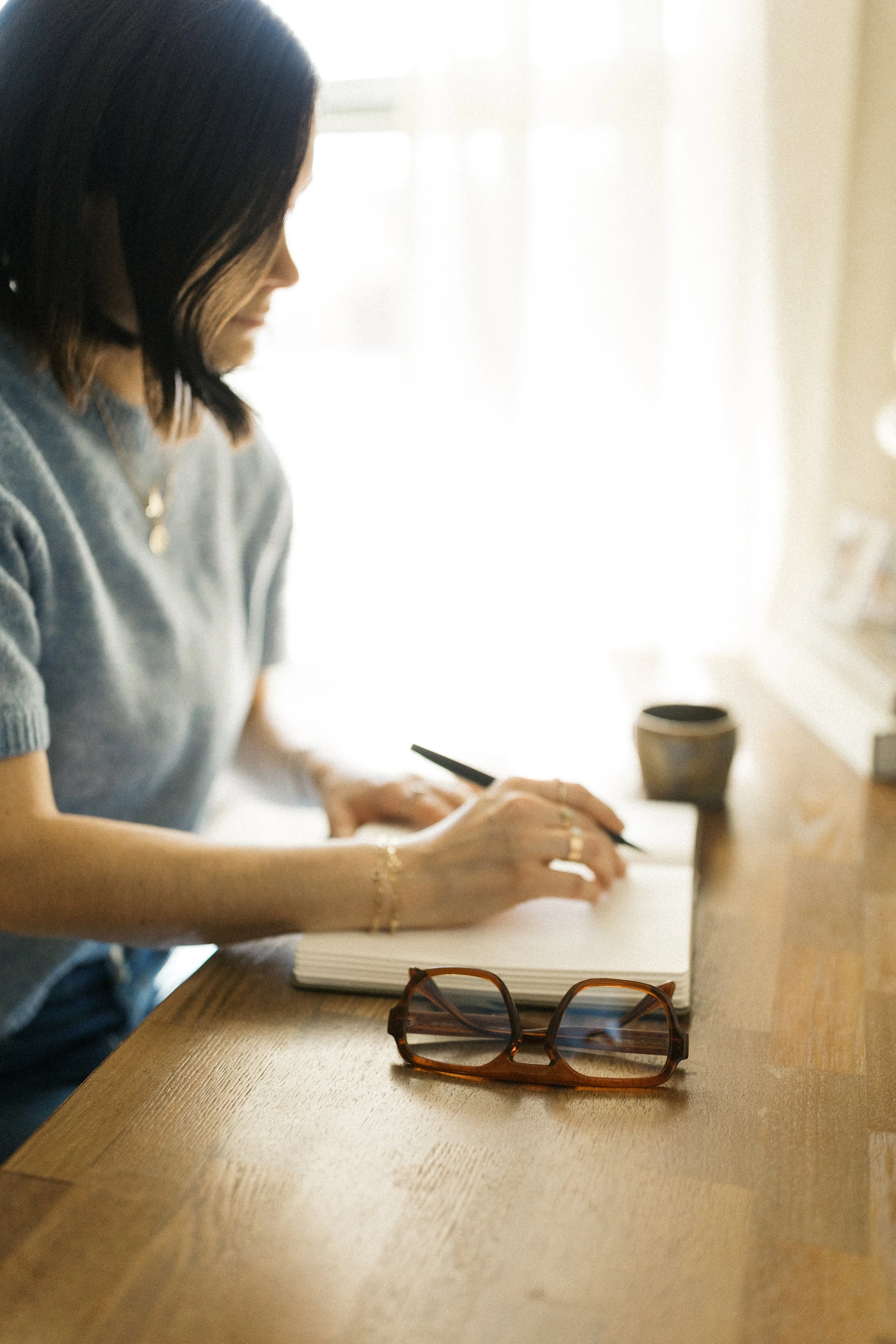 A woman sitting at a wooden table, writing or drawing in a notebook. She is wearing a gray top, with glasses placed on the table in front of her. There is a cup nearby, and the room is bright with natural light coming through a window.