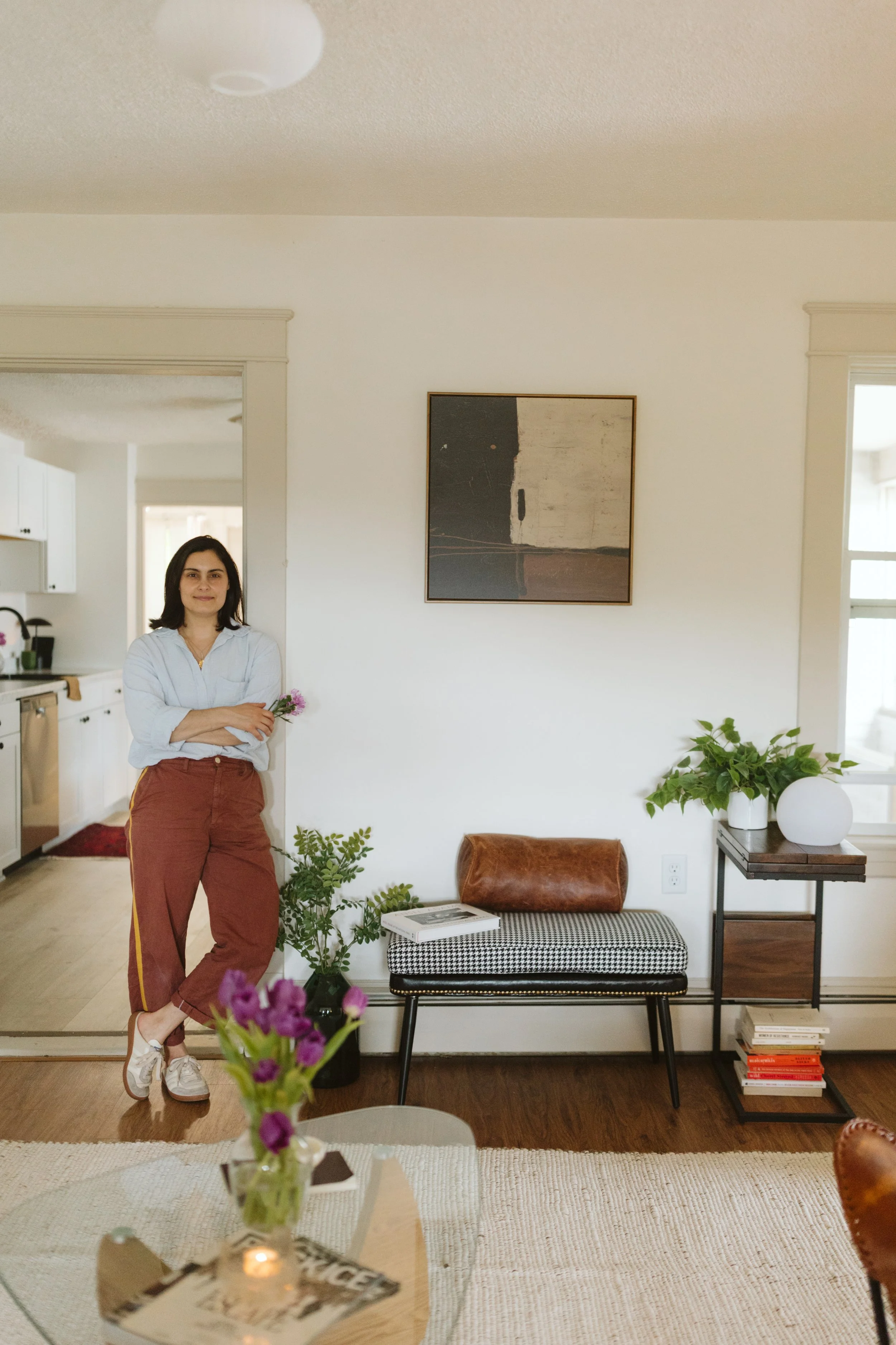 A woman with dark hair, wearing a light blue shirt and brown pants with a yellow stripe, standing in a living room. She is crossed her arms and smiling, holding pink flowers. The room has a white wall with a painting and various plants, furniture, and decor.