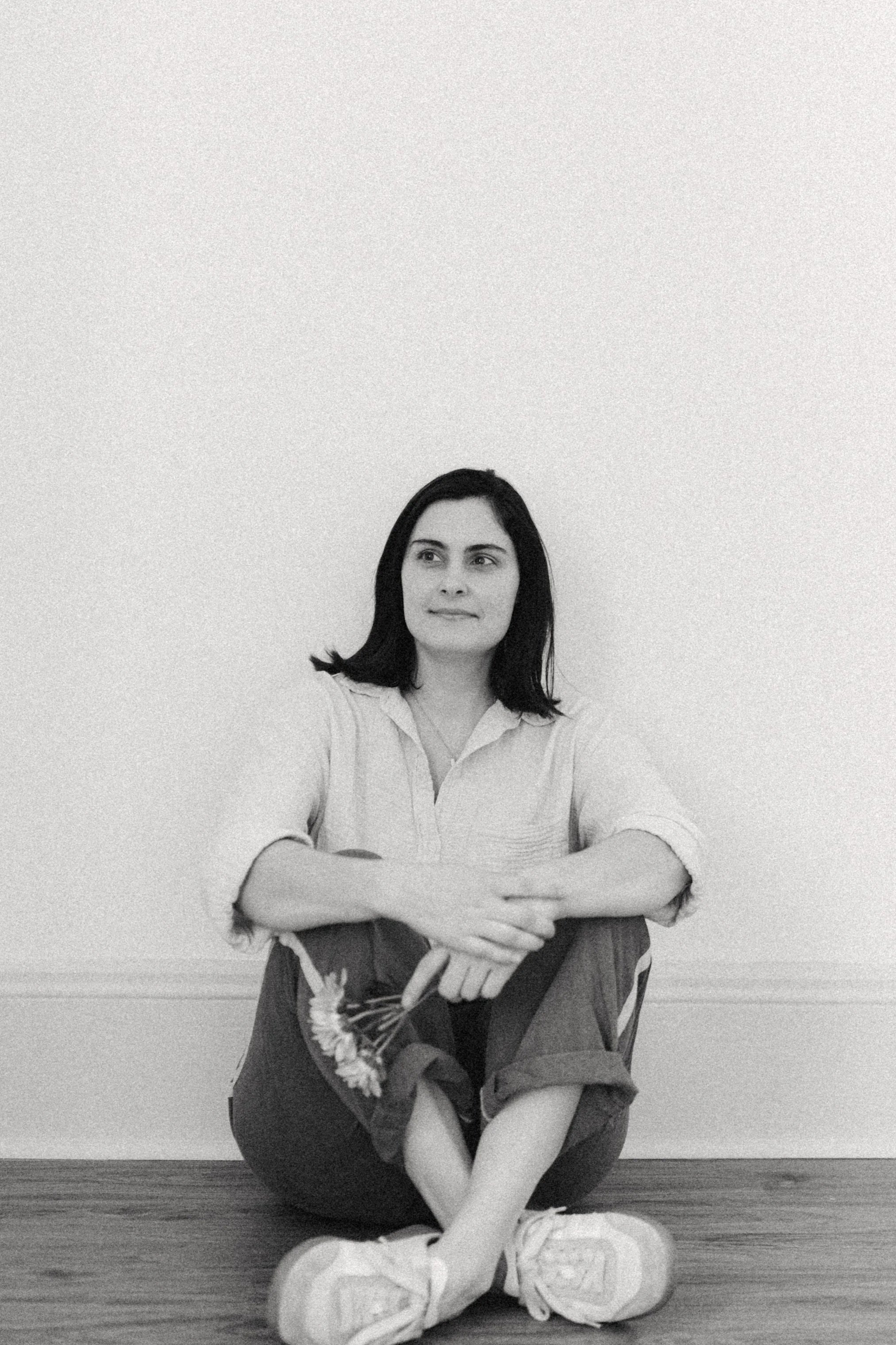 A black and white photo of a young woman sitting on the wooden floor against a plain wall, holding a bunch of flowers and looking off to the side.