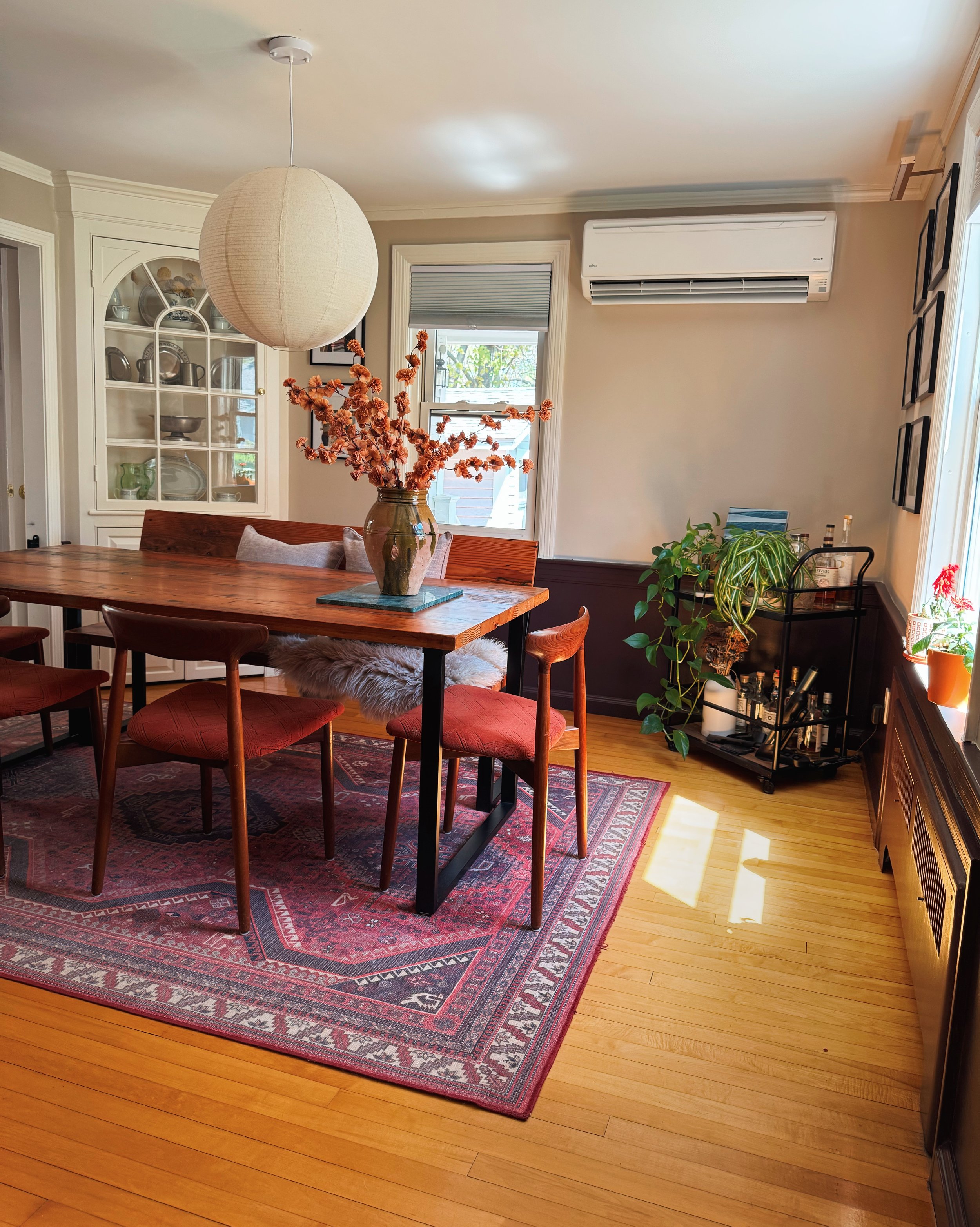 A dining room with wooden table and chairs, a large hanging paper lantern, a decorative rug, a china cabinet, and houseplants near windows with blinds, with sunlight coming through.