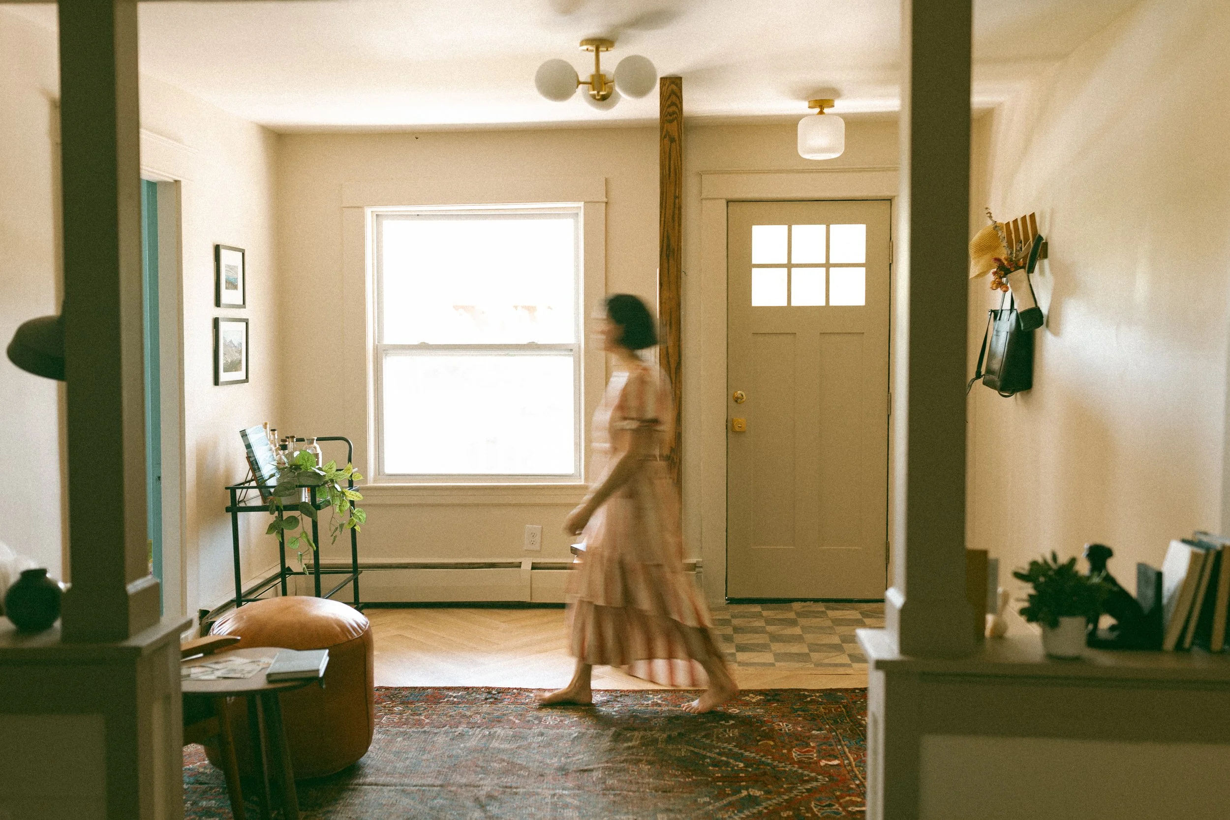 A woman in a pink striped dress walking barefoot inside a cozy living space with natural light from a large window. The room has wooden flooring, a patterned rug, and simple decor, including framed pictures on the wall, a black metal shelf with a plant, and a doorway leading outside.