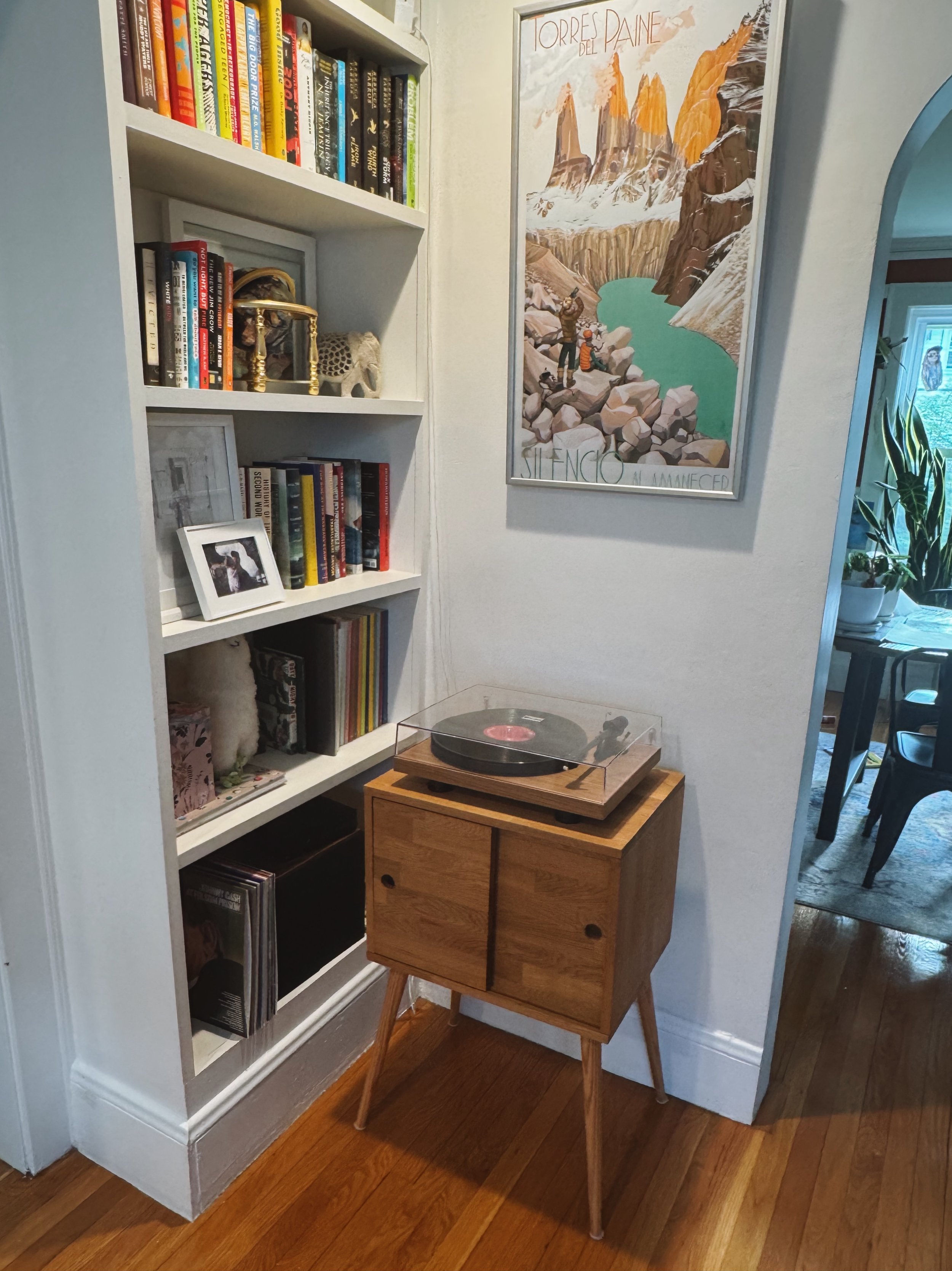 A wooden side table with three drawers is positioned next to a white bookshelf filled with books and some framed pictures. A turntable is on top of the table. A framed Nature poster with the text 'TORRES DEL PAINE' hangs on the wall behind.