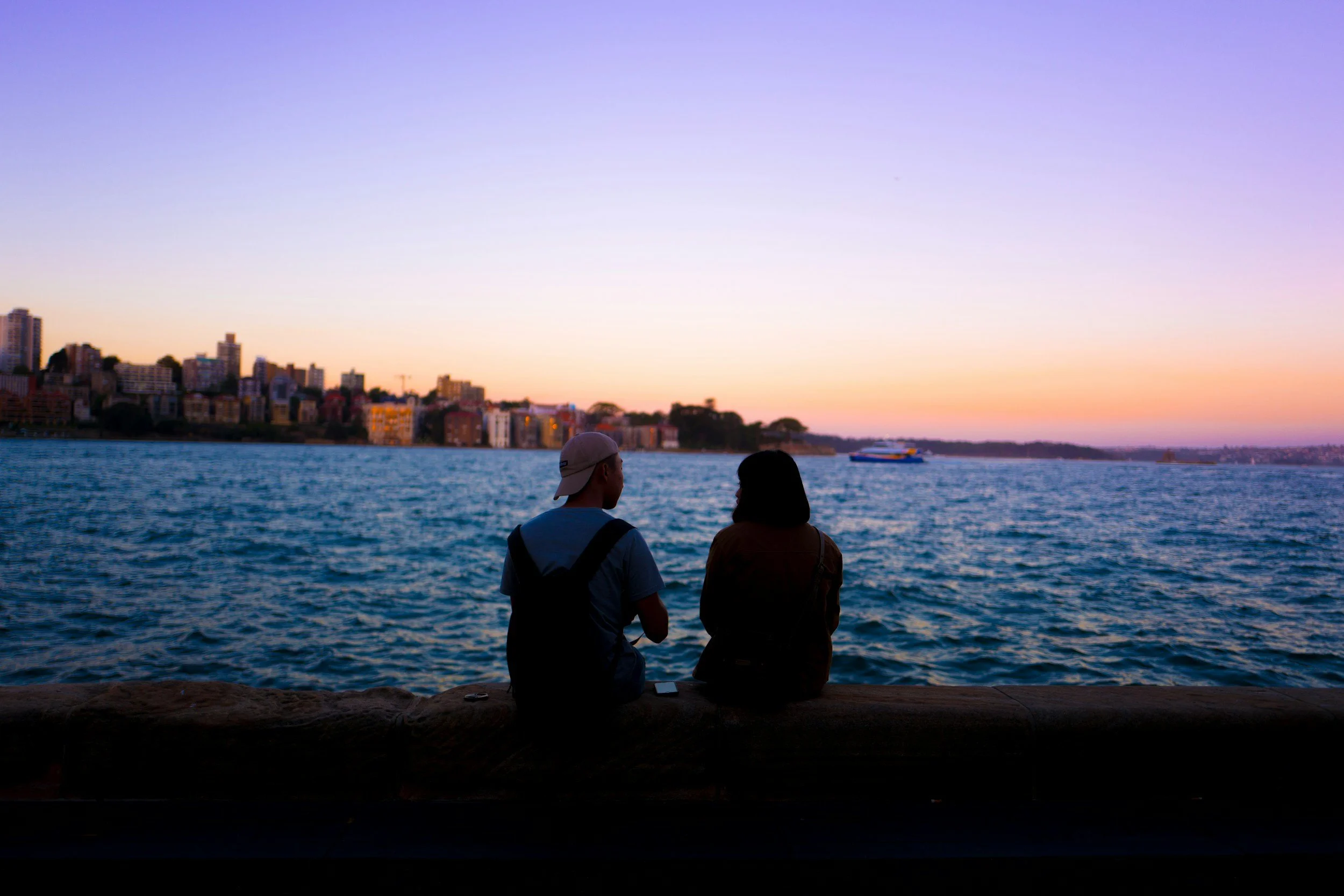 Two people sitting on a stone ledge by the water, watching a sunset with a city skyline in the background, and a boat on the water.