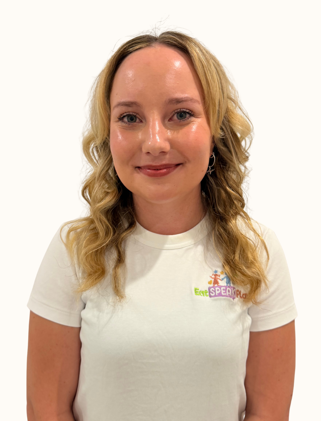 A woman with blonde, curly hair smiling against a plain white background, wearing a white T-shirt with a colorful embroidered logo.