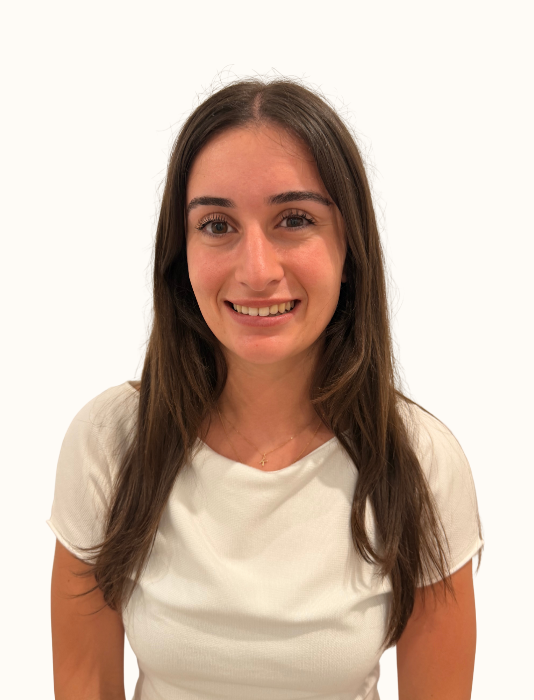 A woman with long brown hair, smiling, wearing a white top and a delicate gold necklace, standing against a plain white background.