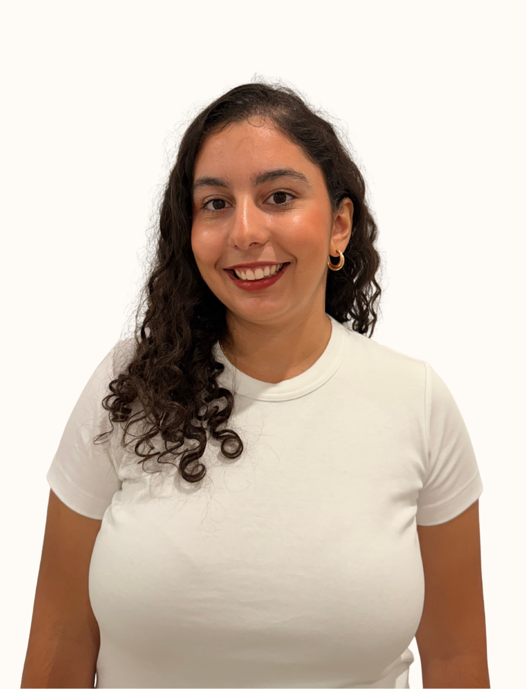 A woman with curly brown hair, wearing a white t-shirt and gold hoop earrings, smiling against a white background.