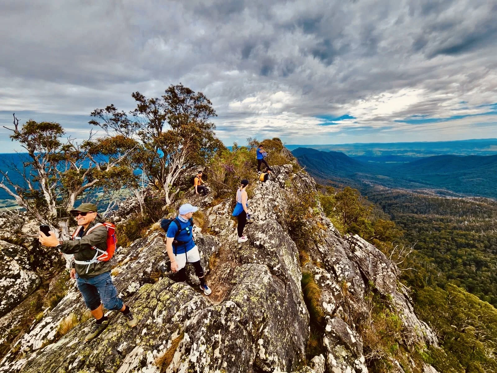 Group of hikers climbing a steep, rocky mountain ridge with a panoramic view of mountains and forest beneath a cloudy sky.