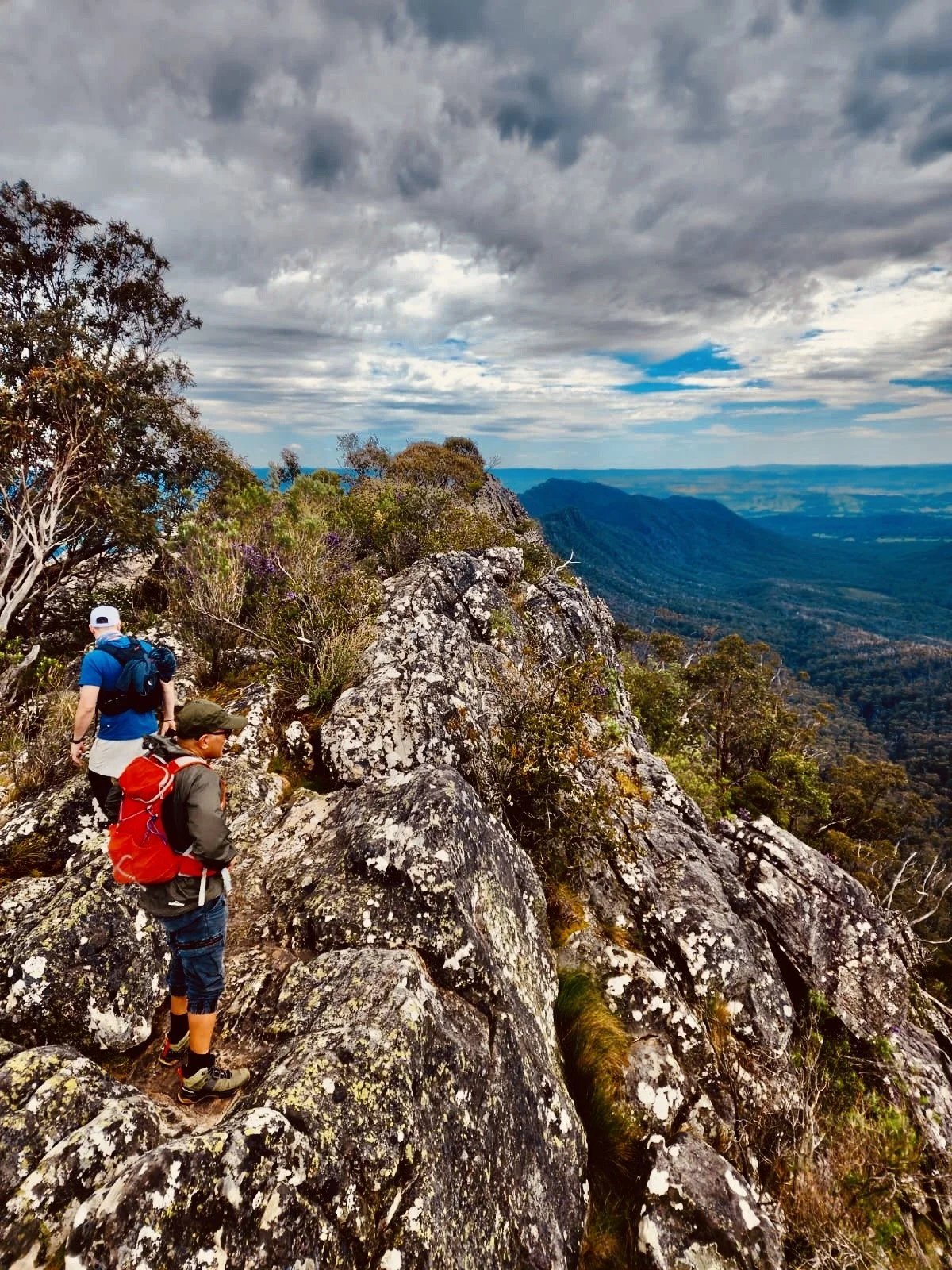 Two hikers with backpacks walking on rocky trail on mountain ridge with scenic valleys and cloudy sky in the background.