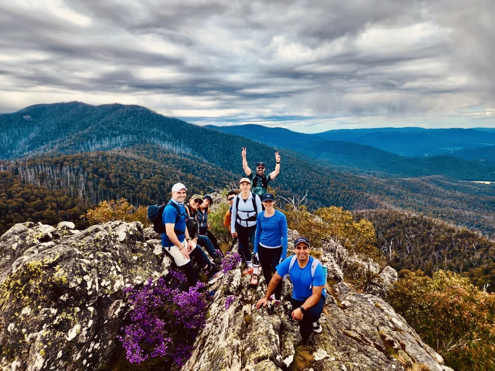 Group of hikers on rocky mountaintop with forested mountains in the background under cloudy sky.
