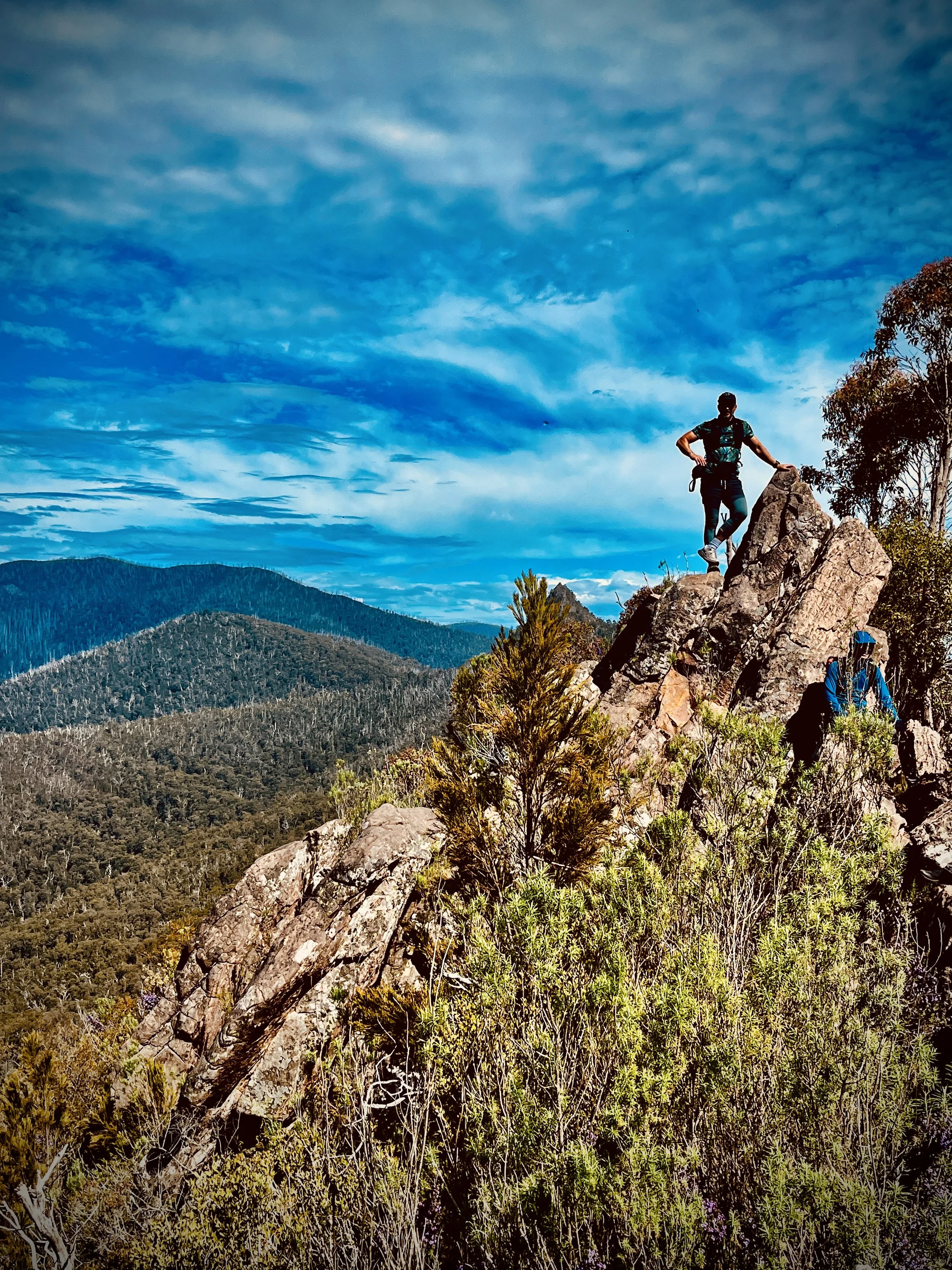 Two hikers standing on large rocks on a mountain trail with a forested valley below and a partly cloudy sky above.