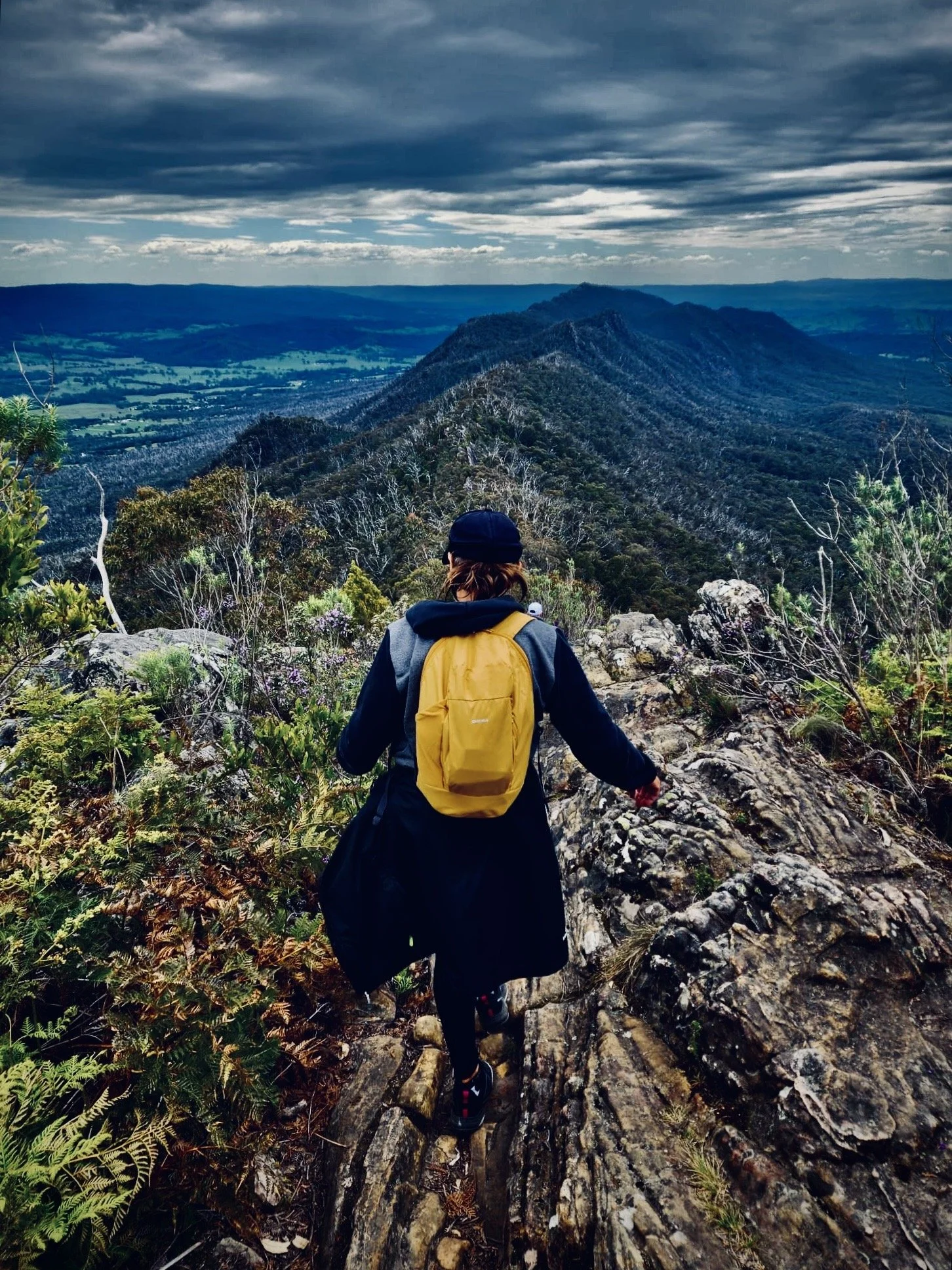 Person hiking on rocky trail, wearing black jacket, black leggings, and a yellow backpack, overlooking a mountainous landscape with forested ridges under a cloudy sky.