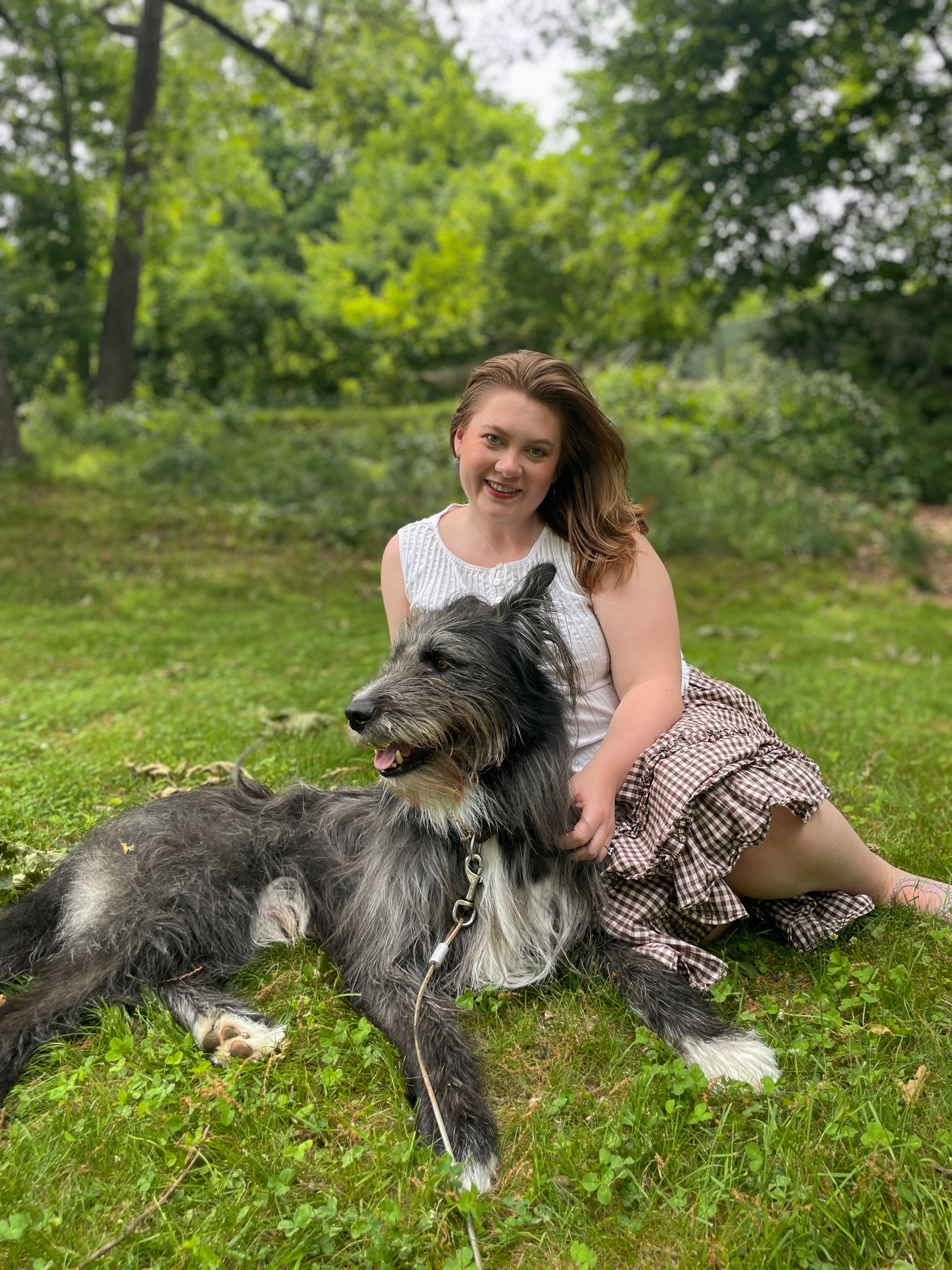 A woman with light skin and long, wavy strawberry blonde hair sitting on the grass outdoors, holding a large, fluffy, gray and black dog. They are in a lush green park with trees in the background. The woman is smiling at the camera.