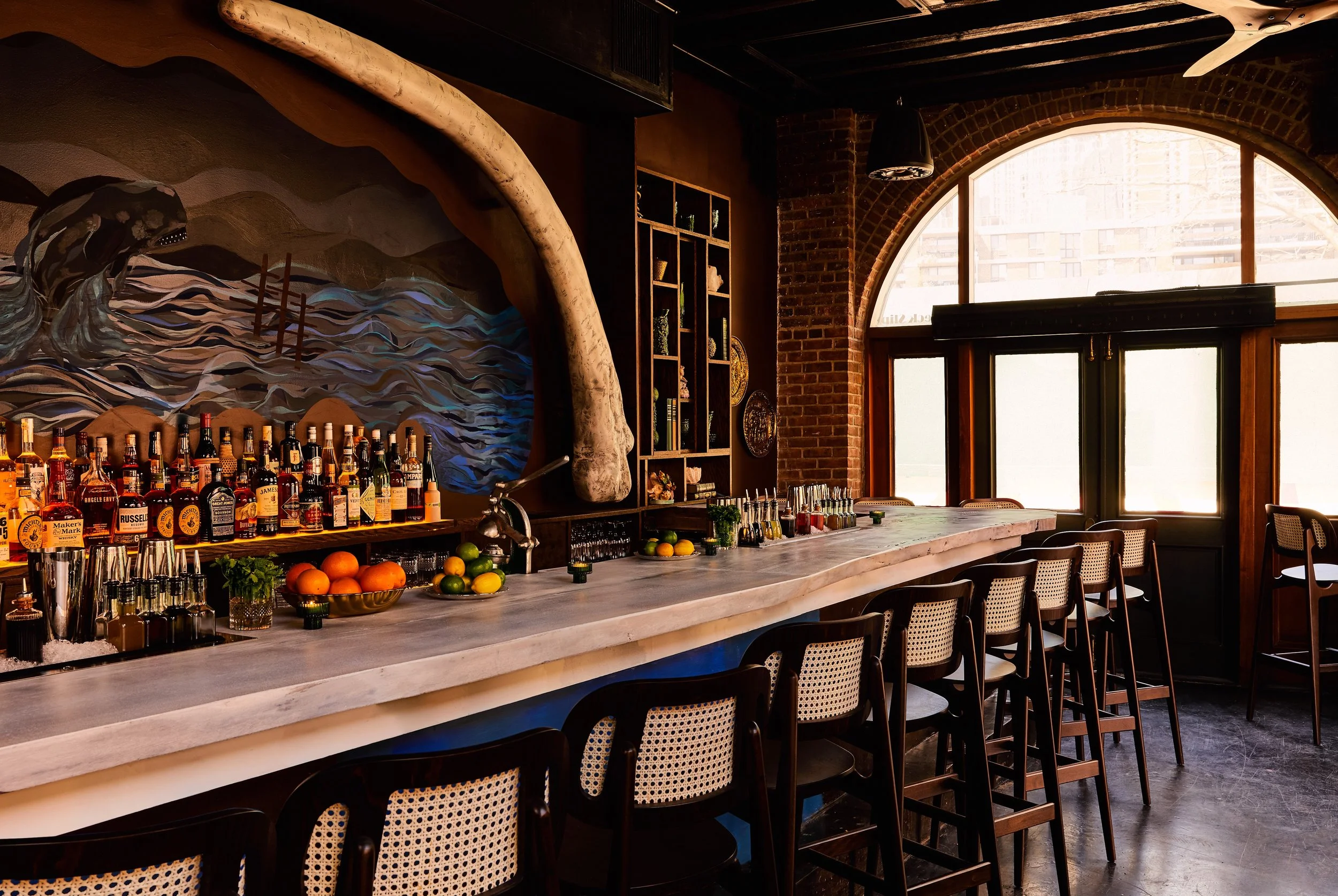 A small round table with a single lit candle on it, flanked by two chairs with woven backrests, in front of a brick wall with a large framed painting of an orange octopus swimming in blue water.