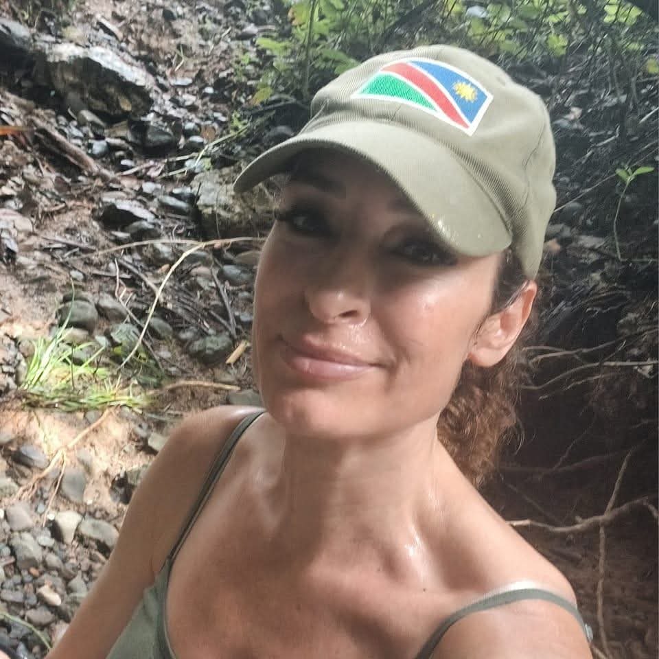 A woman wearing a beige cap with a colorful patch featuring the Philippine flag is smiling in a natural outdoor setting with rocks and greenery.