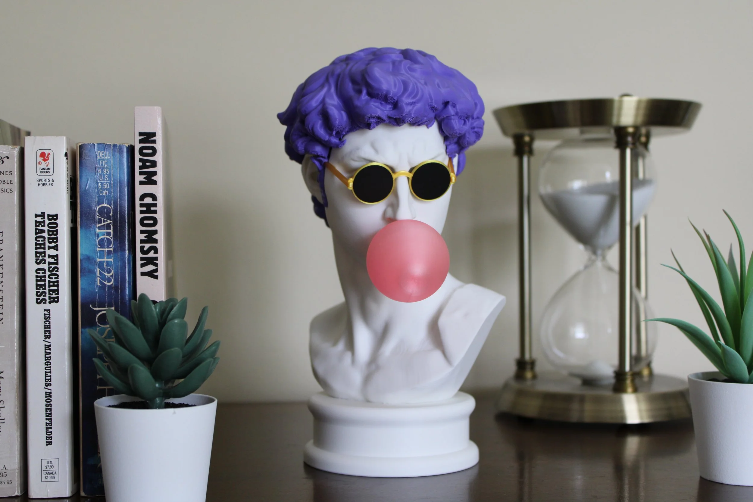 Decorative bust of a woman with purple hair, black sunglasses, and a pink bubblegum bubble, flanked by potted succulents, and an hourglass on a wooden surface.