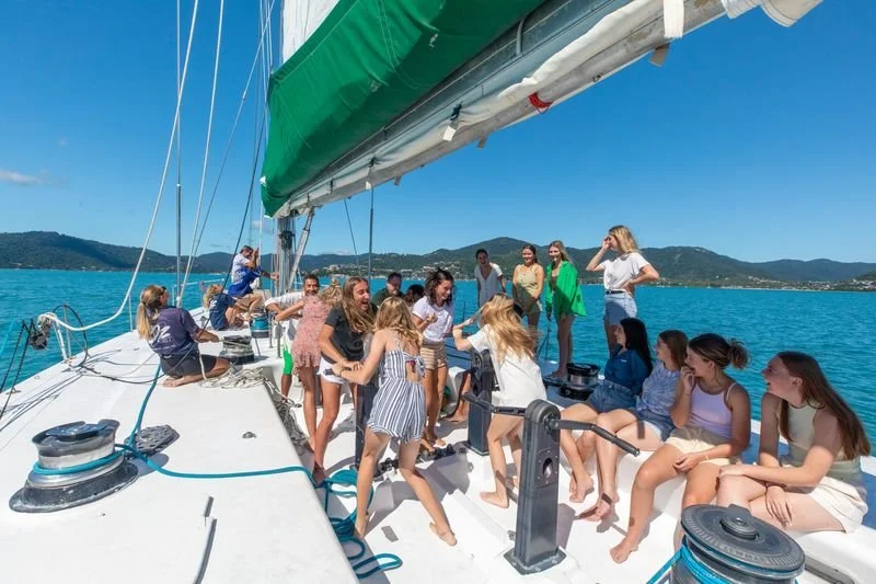 Group of young people enjoying a boat trip on a large sailboat on a sunny day with blue skies, surrounded by calm water and distant hills.