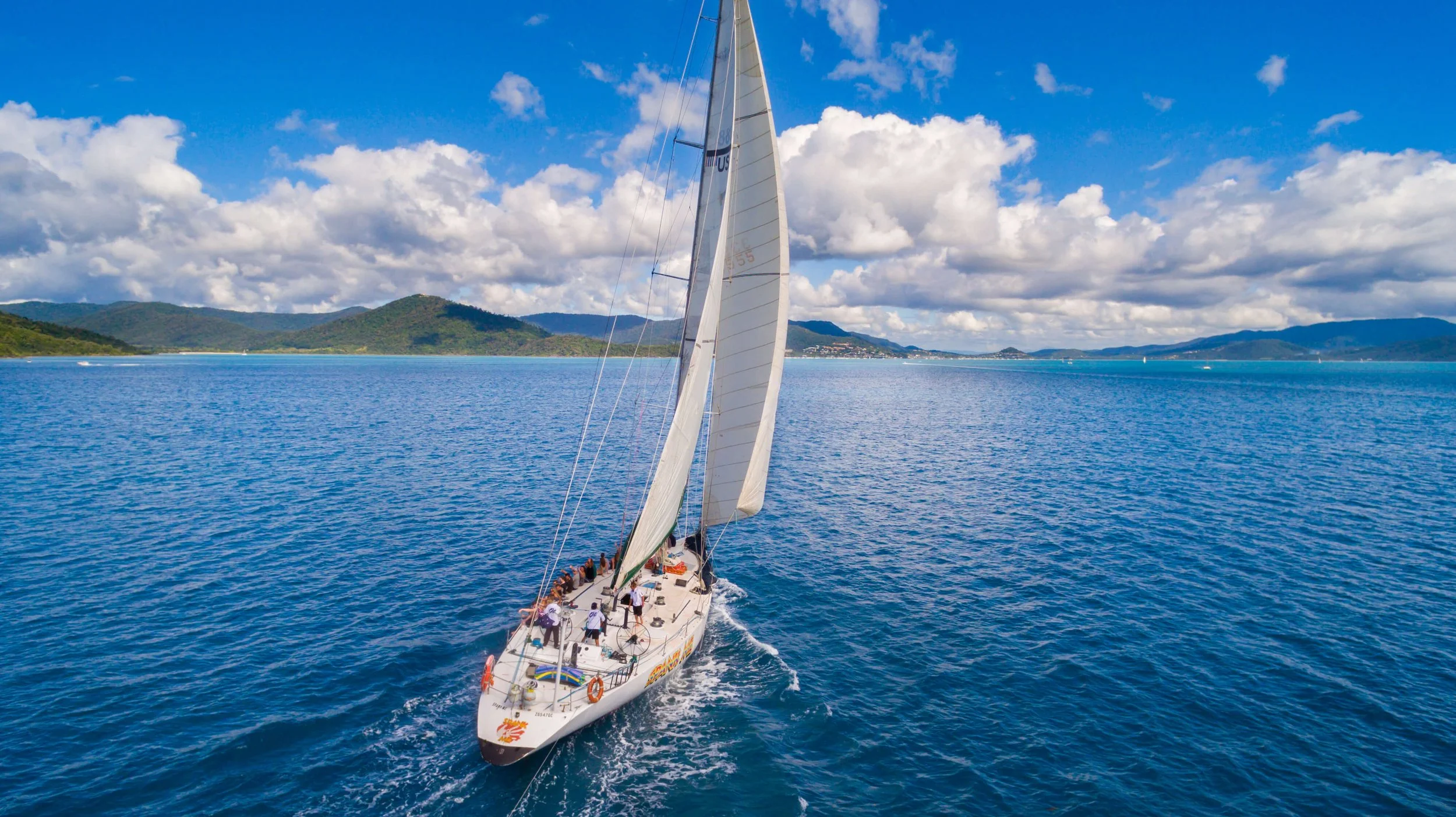 A sailboat with white sails sailing on blue ocean water, with land and green hills in the background under a partly cloudy sky.