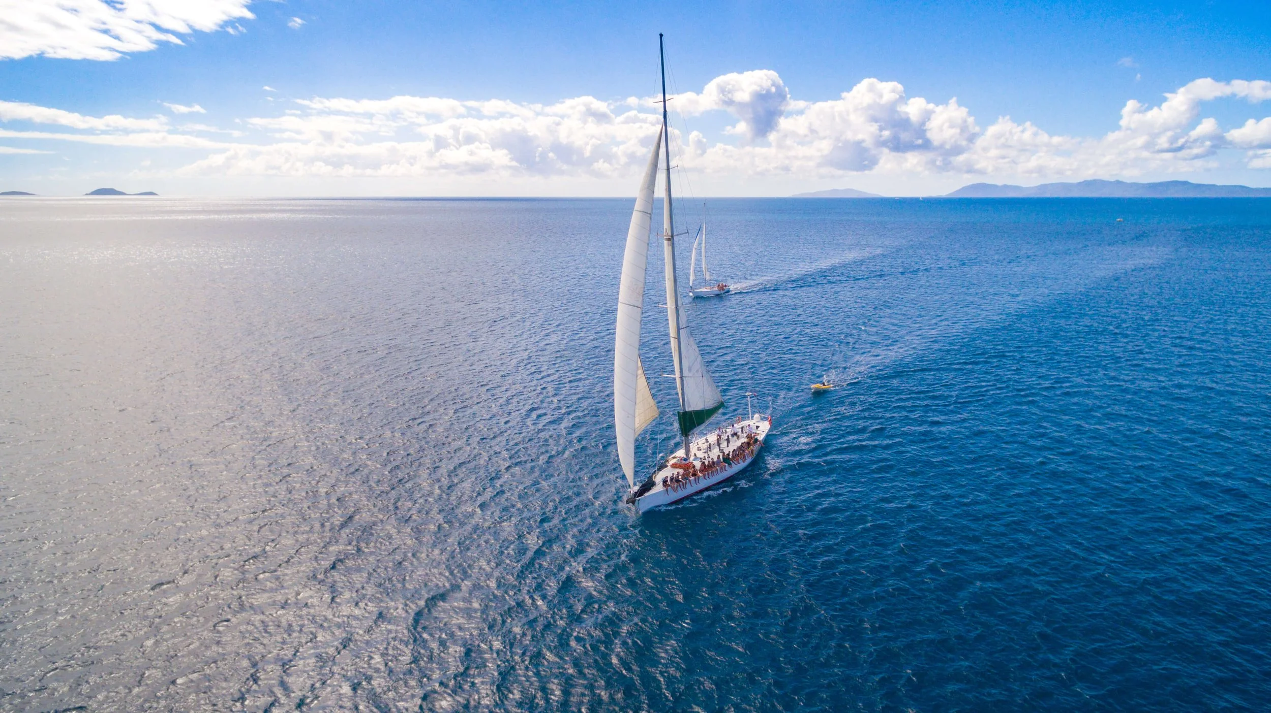 Sailboat with people on deck sailing in open blue ocean with distant islands and a partly cloudy sky.