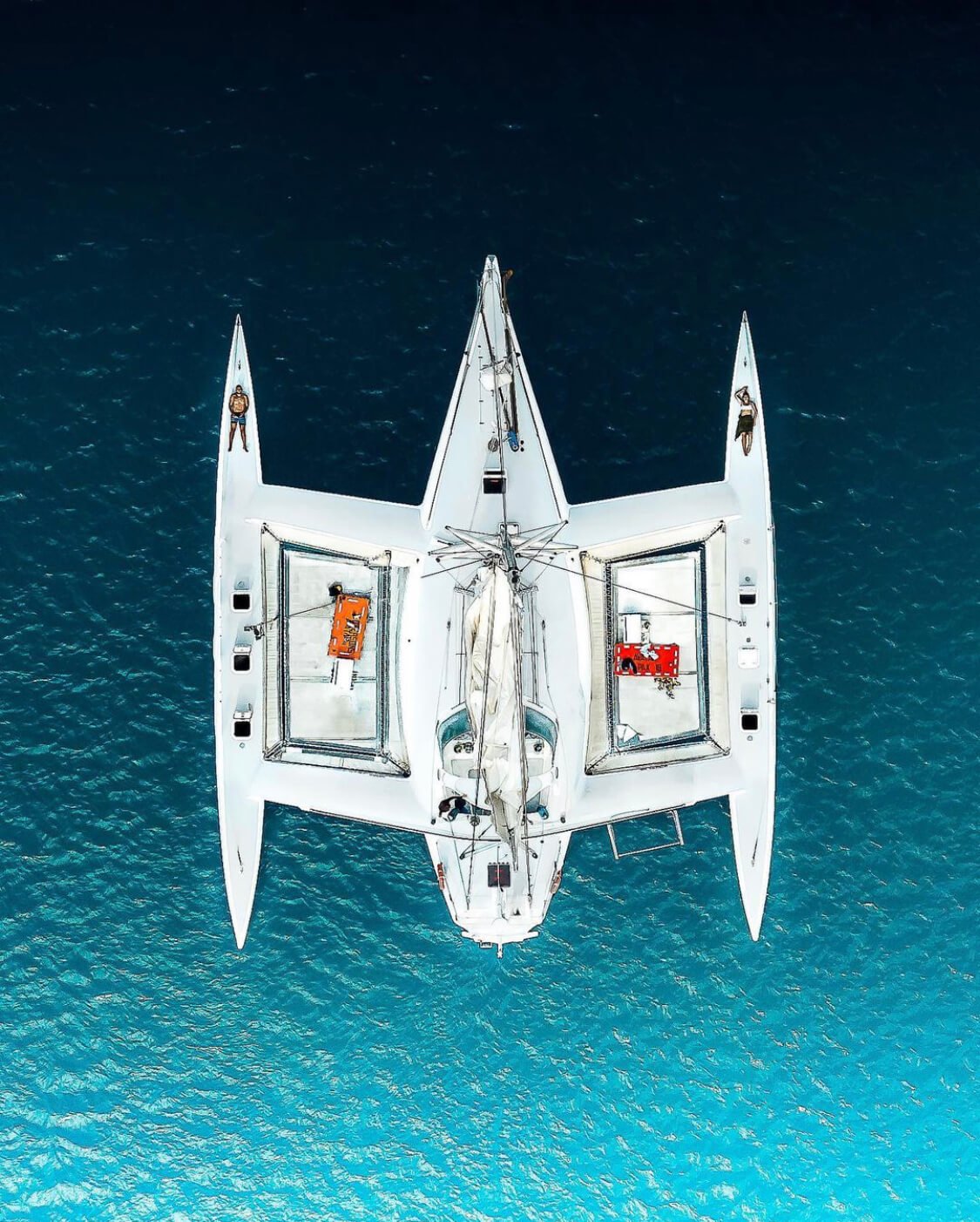 An aerial view of a white yacht on blue ocean water, with two people sunbathing on the deck and various equipment and storage boxes visible.