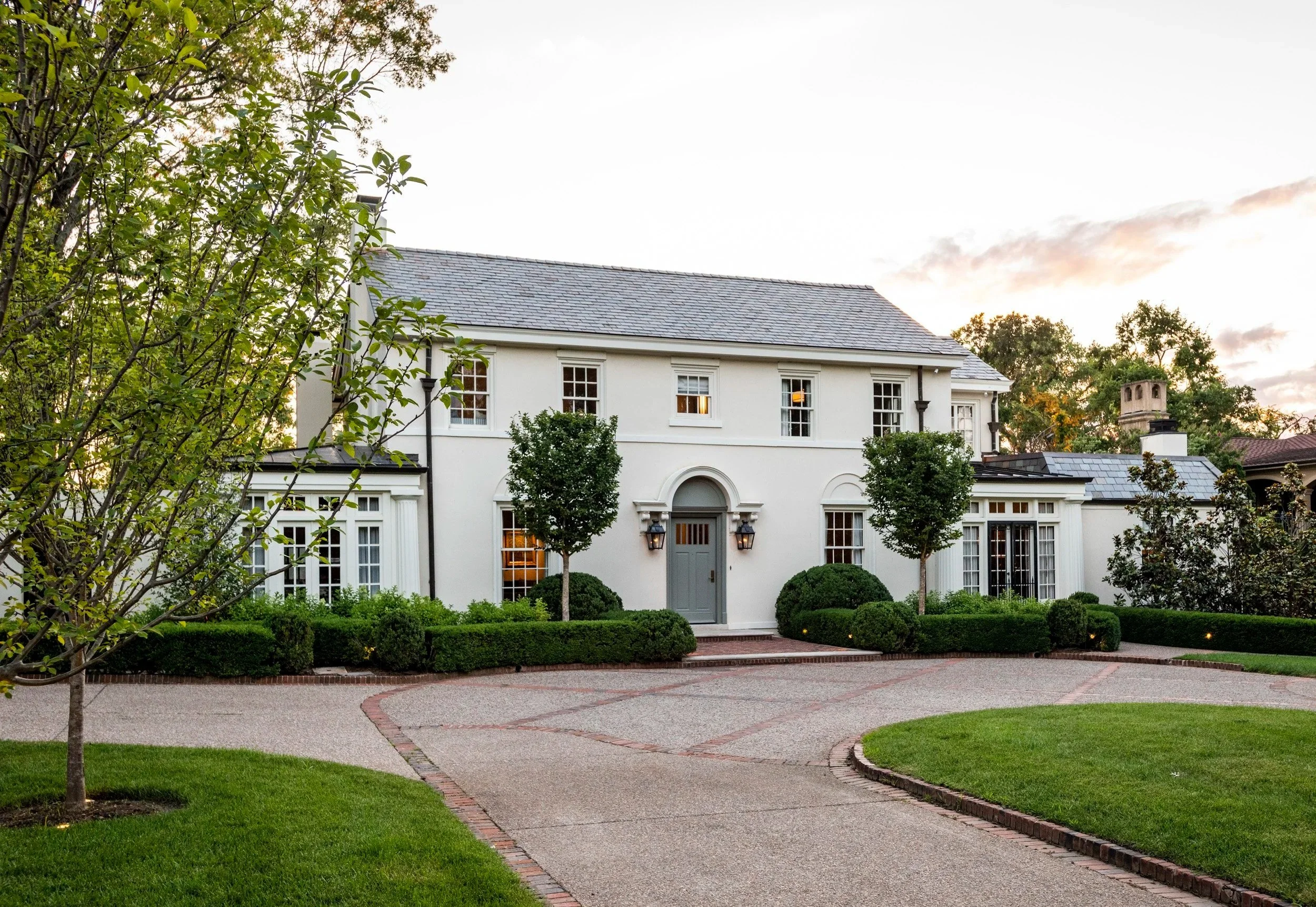 A large white house with multiple windows and a curved front door surrounded by neatly trimmed bushes and a small tree. The house has a gray shingle roof and is set against a background of trees and a partly cloudy sky during sunset. The front yard features a paved circular driveway with a brick border, and the grass is lush and green.
