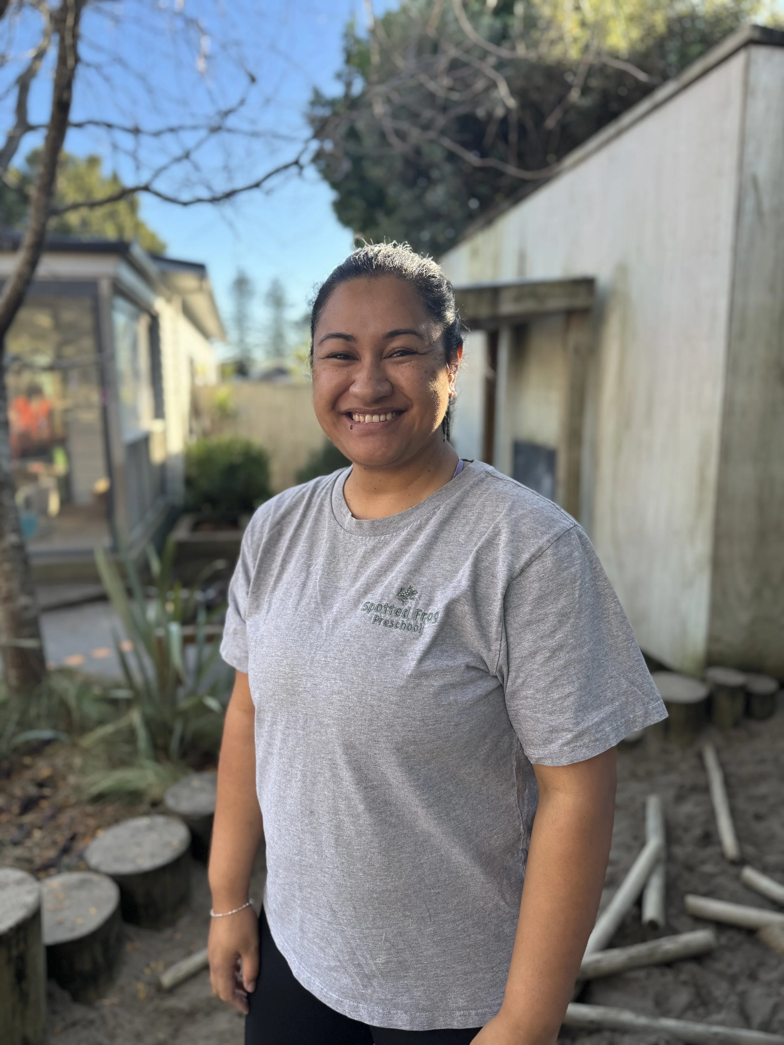 Female ECE teacher smiling with playground background and grey tshirt with Spotted Frog Logo