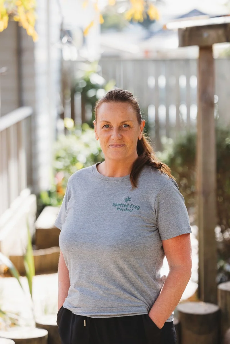 A female ECE teacher with brown hair in a ponytail standing outdoors, wearing a grey t-shirt with green text that says 'Spotted Frog Preschool' and black pants, smiling gently at the camera.