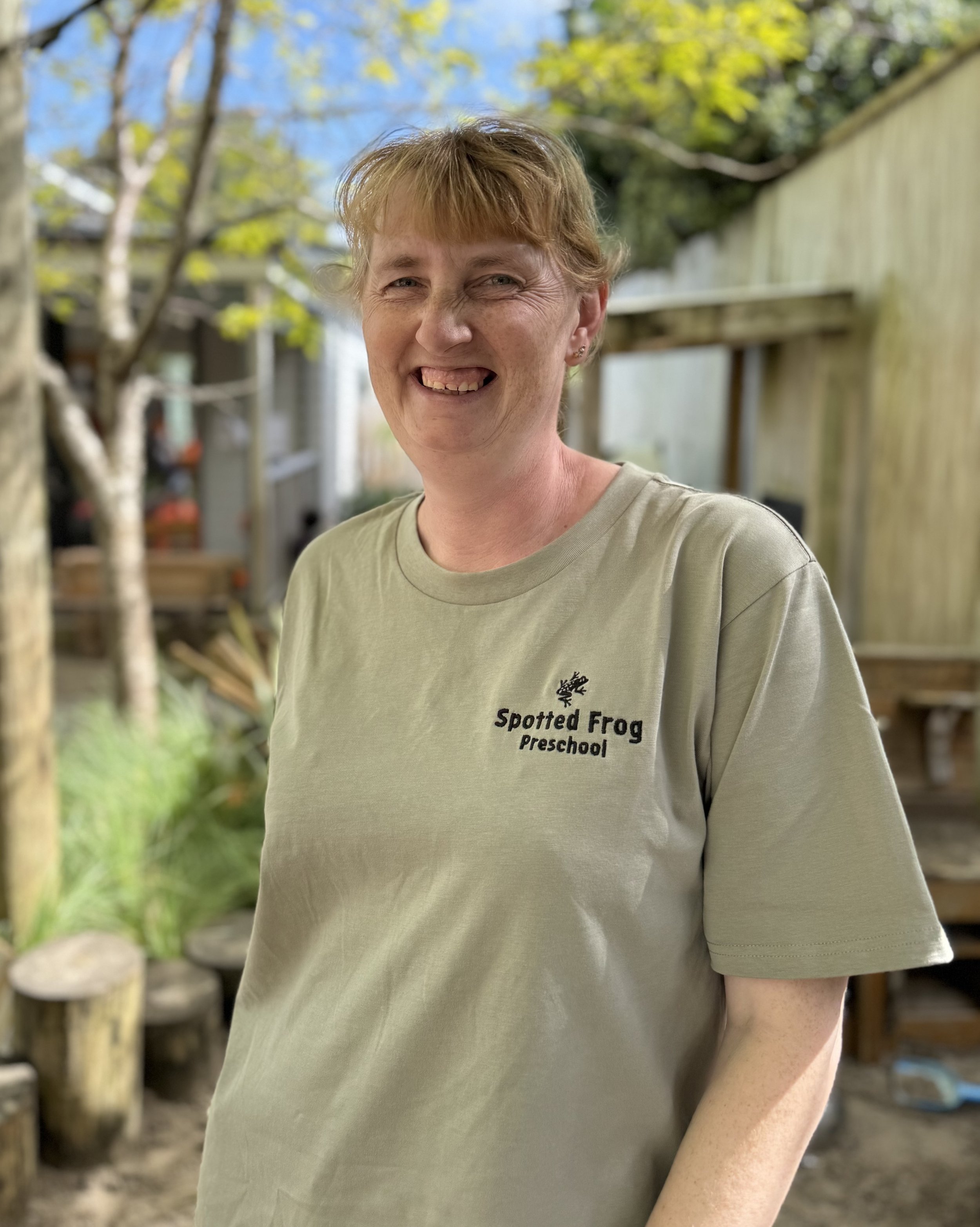 Smiling qualified ECE teacher in a Spotted Frog t-shirt, learning against a wooden post in the preschool outdoor playground.